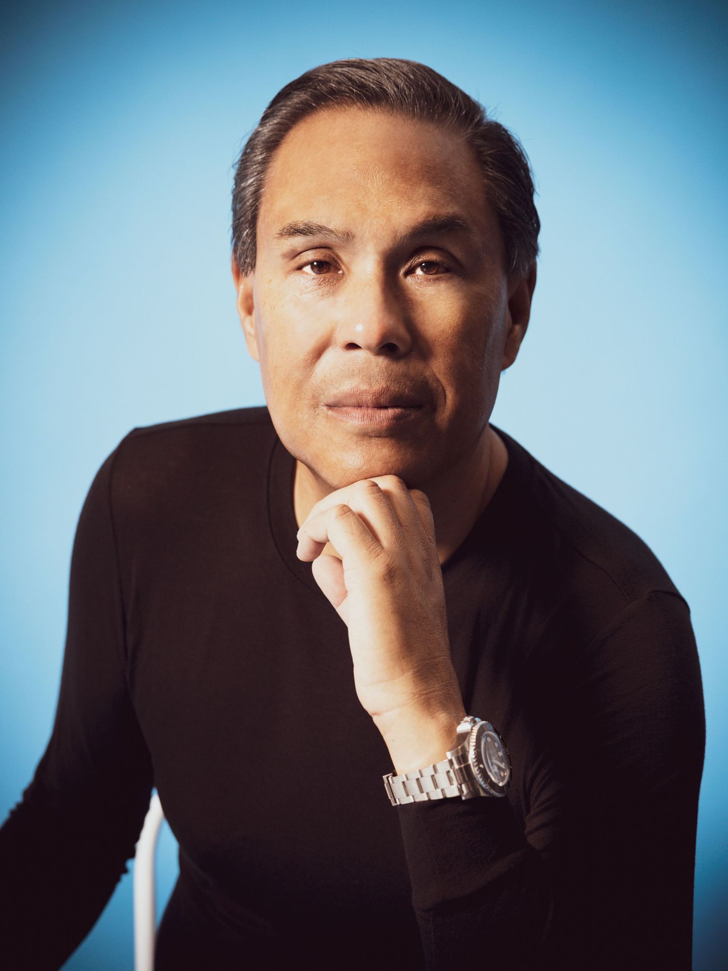 Professional man in black turtleneck poses thoughtfully with hand on chin against blue background.