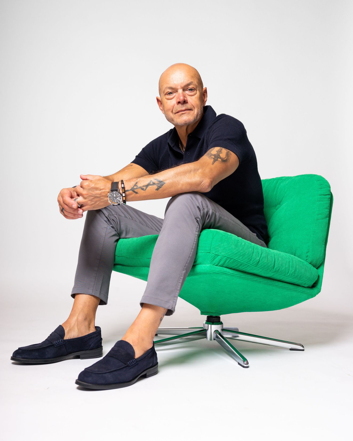 Bald man with tattoos sits relaxed in a green modern chair, wearing gray pants, navy polo, and blue loafers.