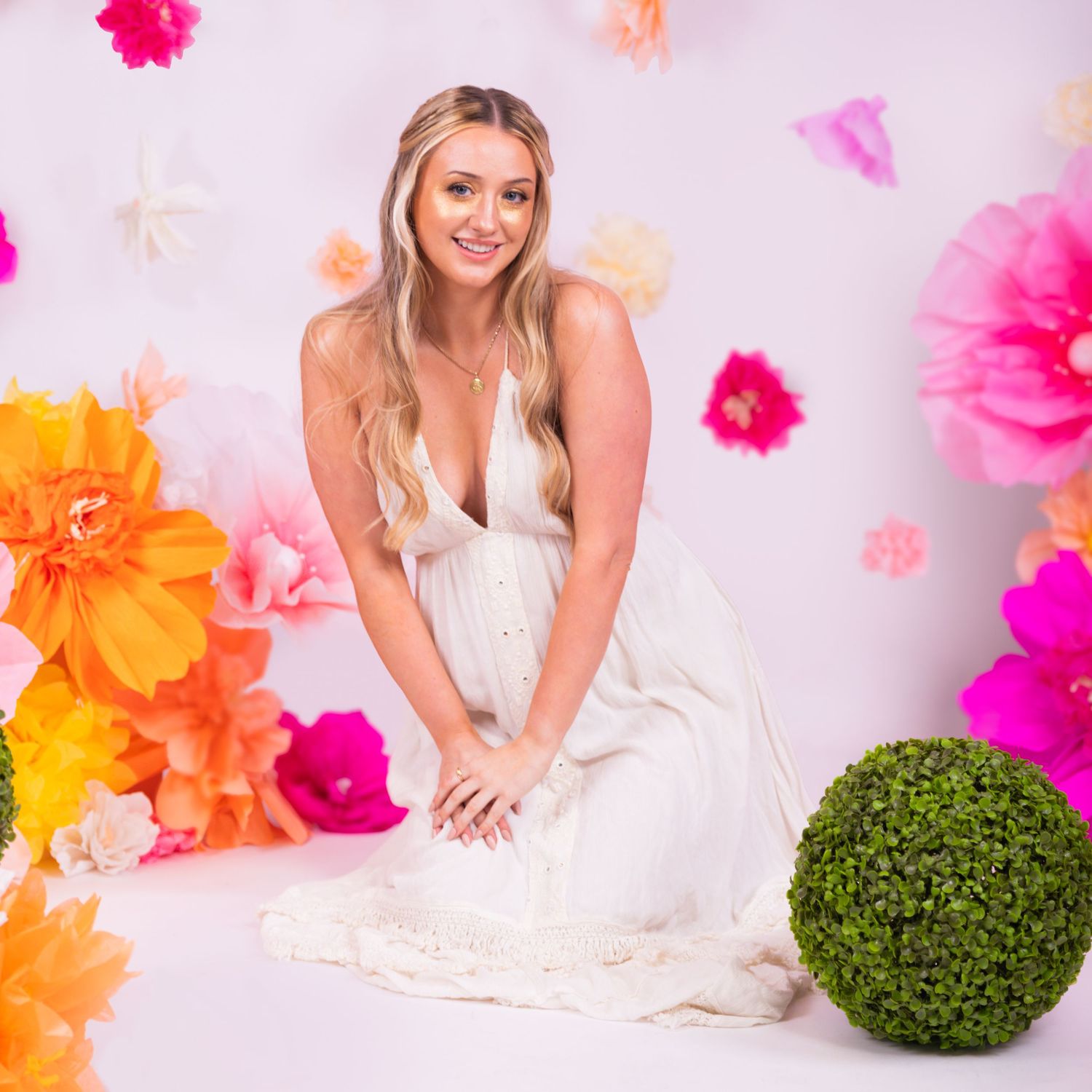 Young blonde woman in white dress poses gracefully among colorful paper flowers in a bright studio setting.