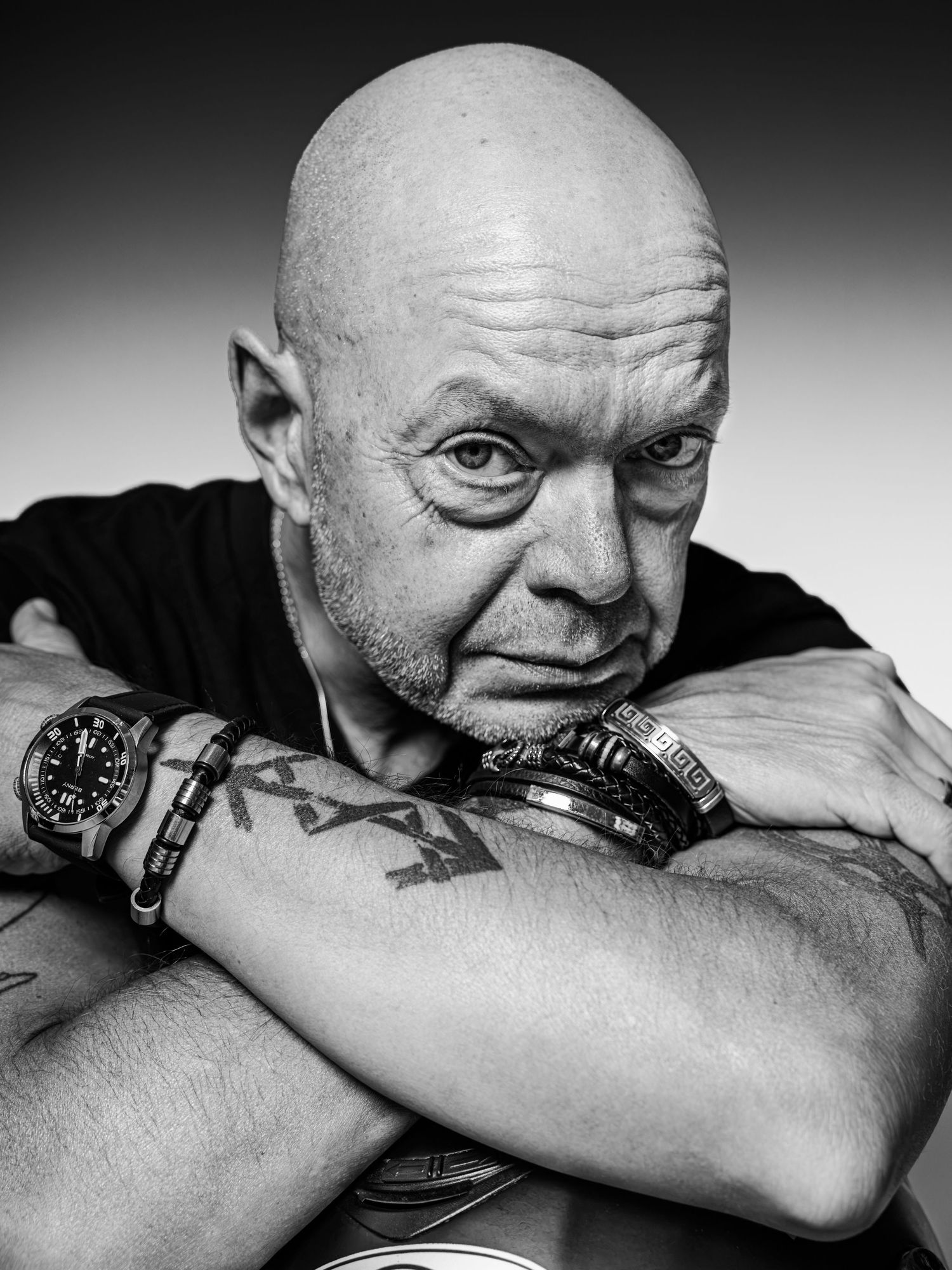 Black and white portrait of a bald tattooed man with crossed arms wearing watches, intense gaze.