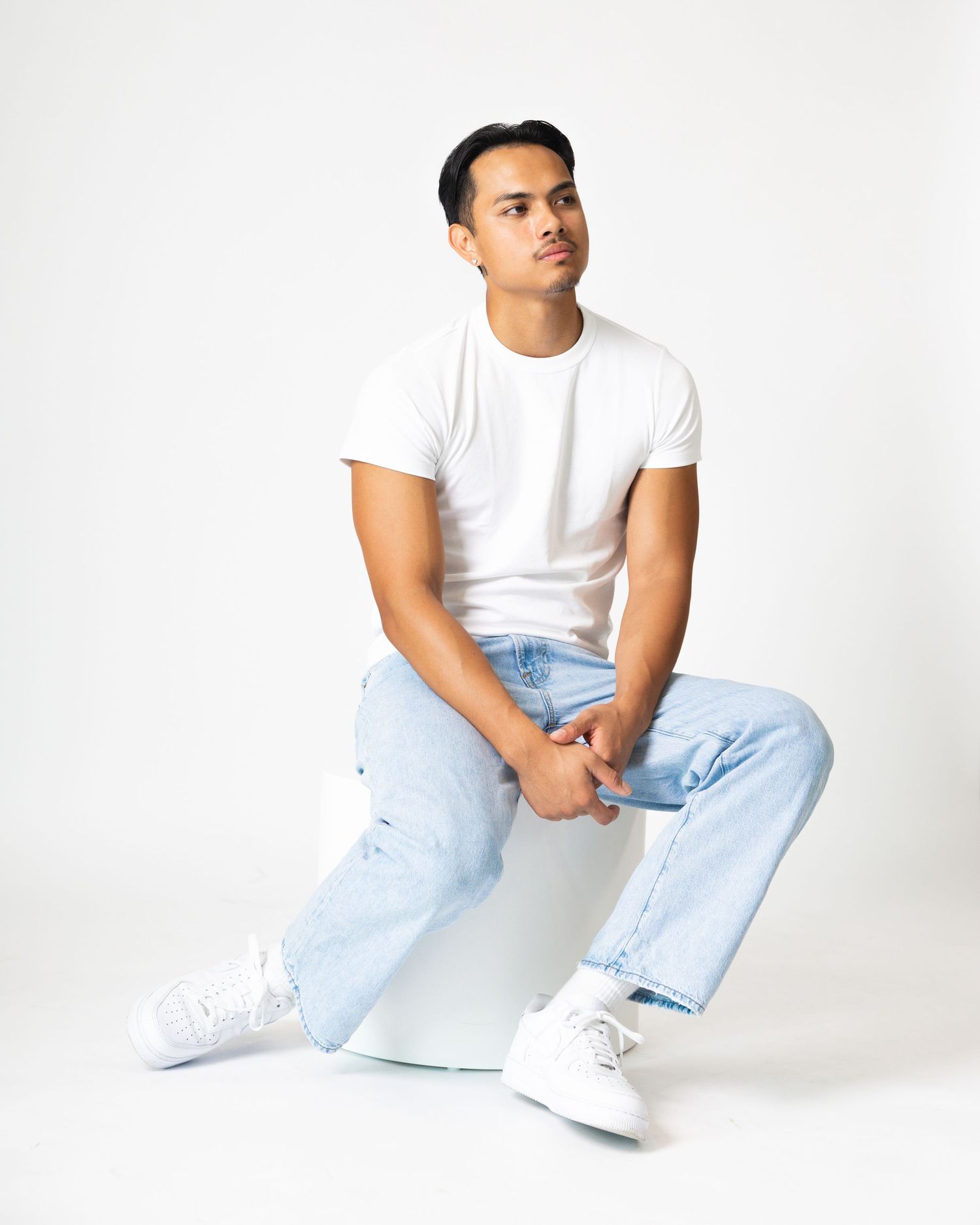 Young man in white t-shirt and light blue jeans sitting casually on a white cube against a white background.