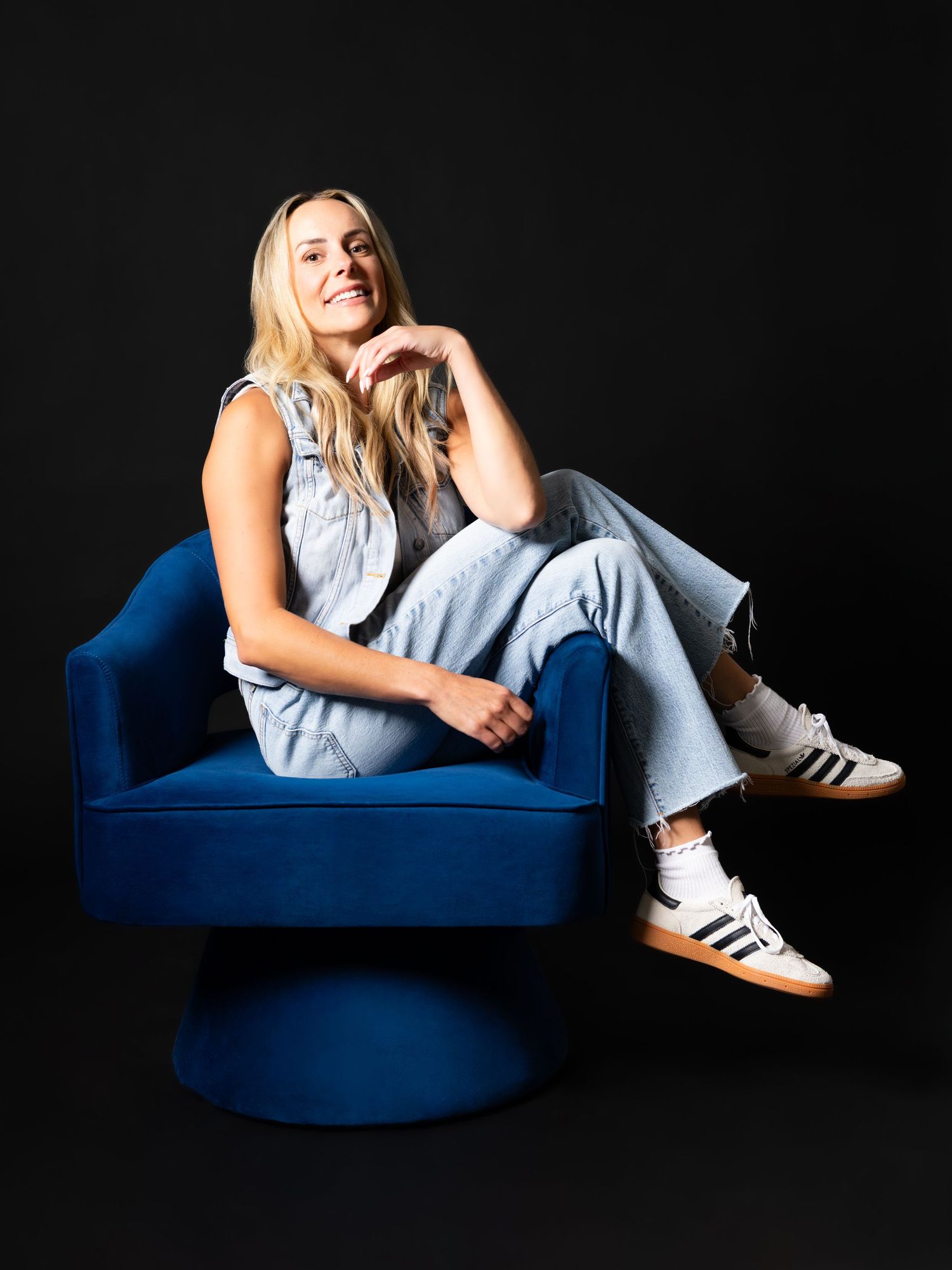 Blonde woman sitting relaxed in a blue velvet chair wearing denim overalls and Adidas sneakers.