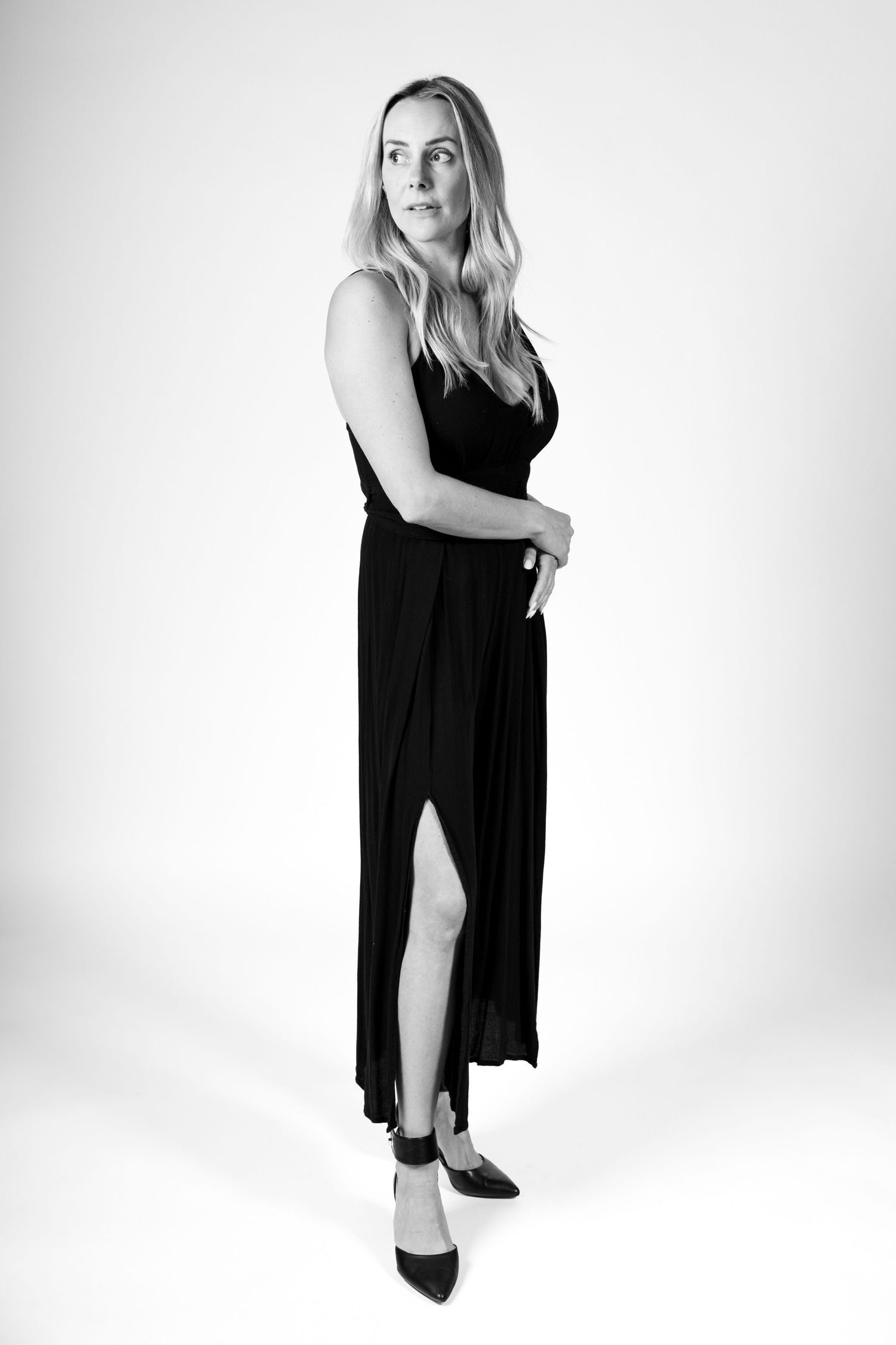 Black and white photo of a blonde woman in an elegant black maxi dress with slit, posing in heels.
