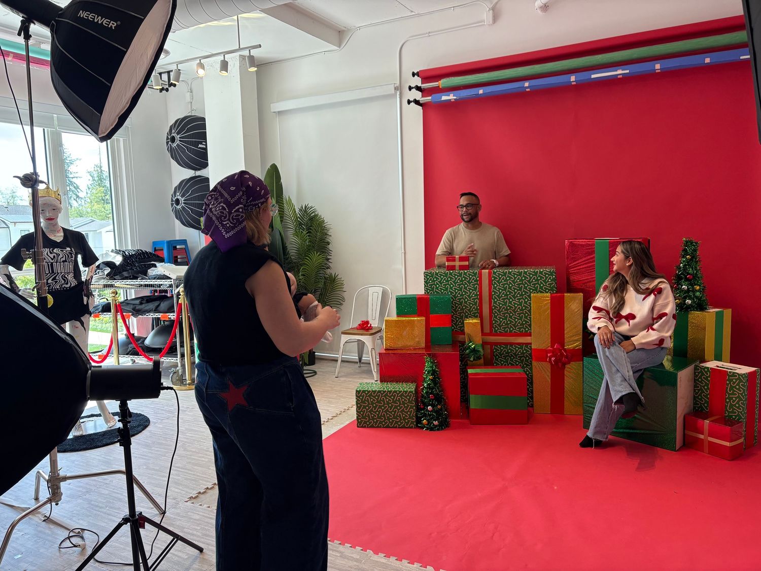 Behind-the-scenes Christmas photo shoot with colorful gift boxes, red backdrop, and festive holiday decorations in a studio.