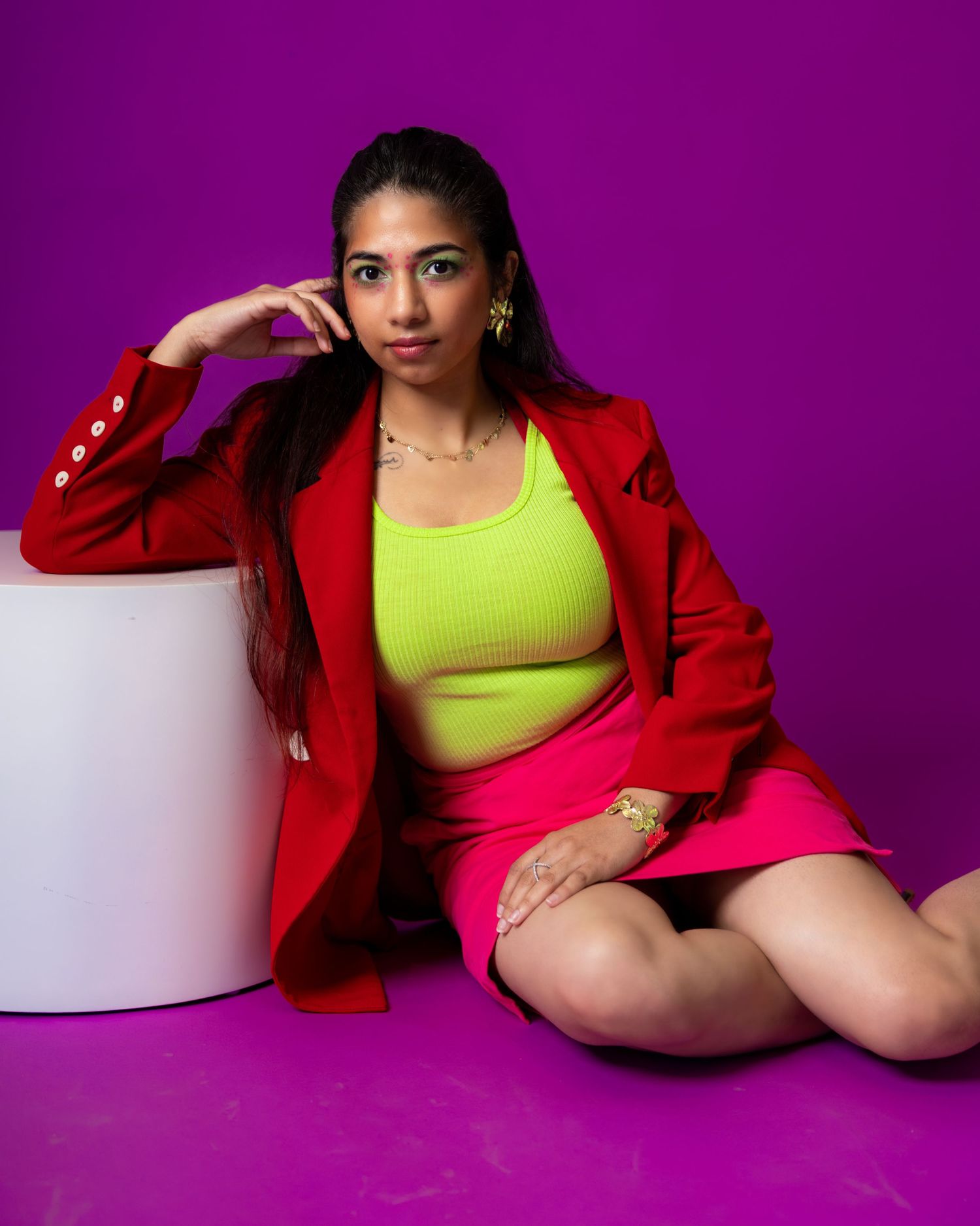 Woman in red blazer, neon yellow crop top, and pink skirt poses against a vibrant purple background.