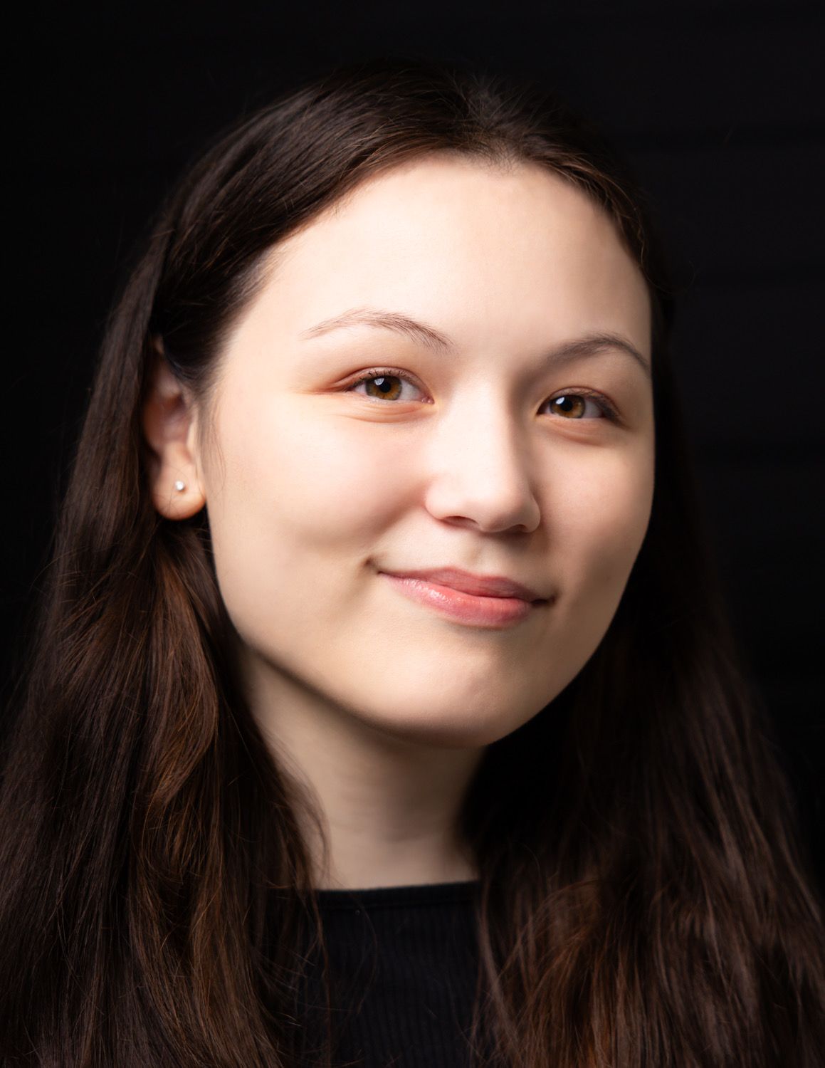 Young woman with long dark hair smiling warmly against a dark background in a professional headshot photo.