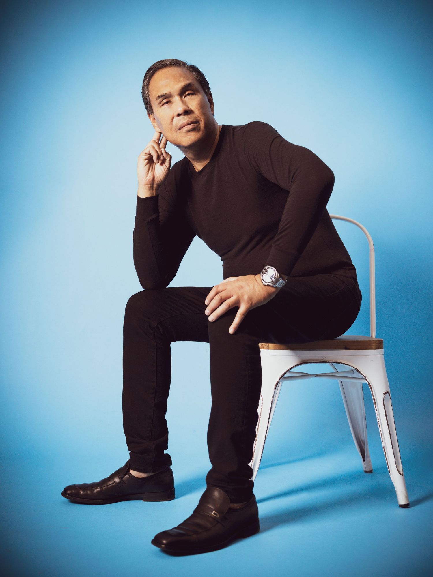 Man in black outfit sits on white chair against blue background, posing thoughtfully with hand near chin.