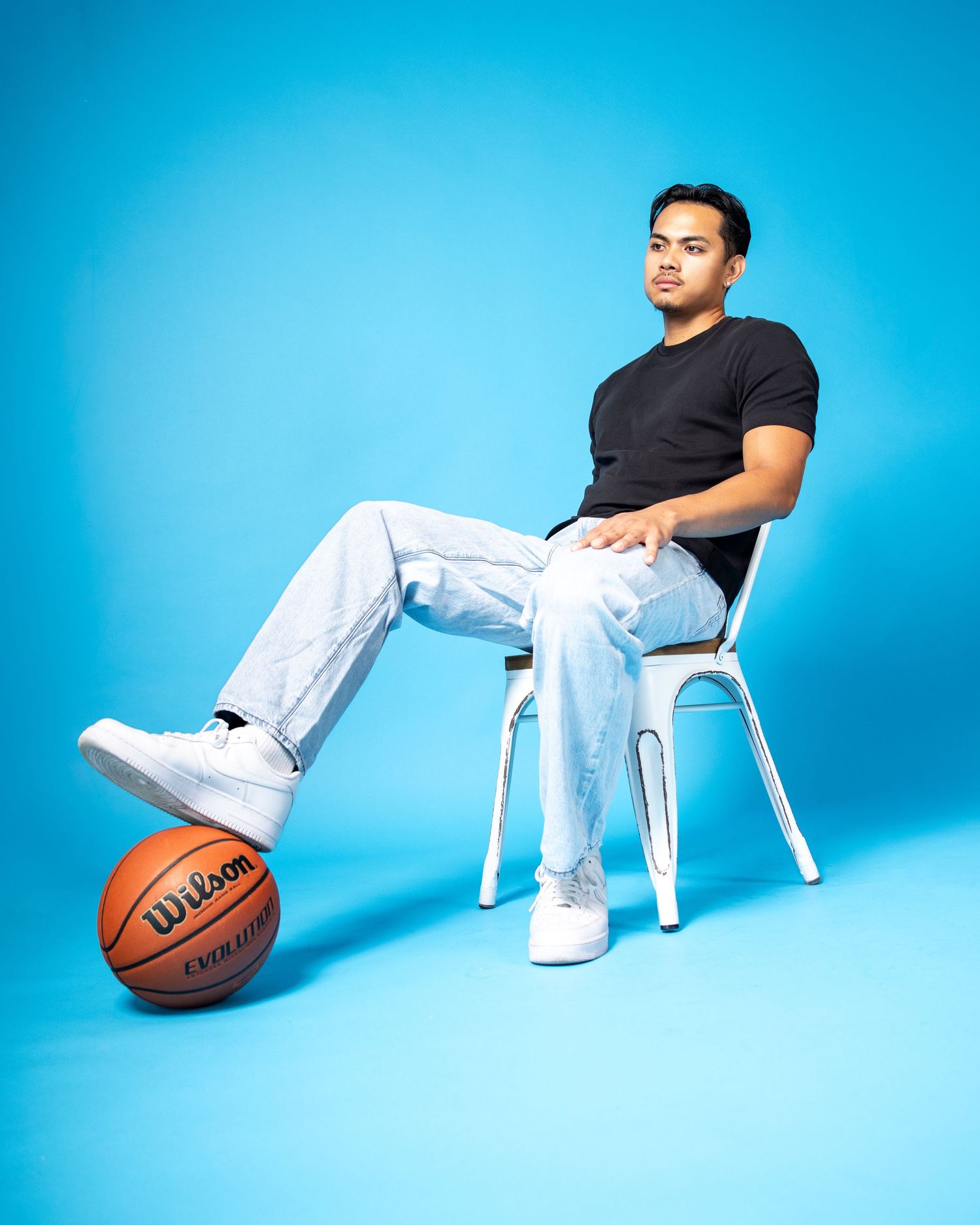 Young man in black t-shirt and light jeans sits on white chair with foot resting on Wilson basketball against blue background.