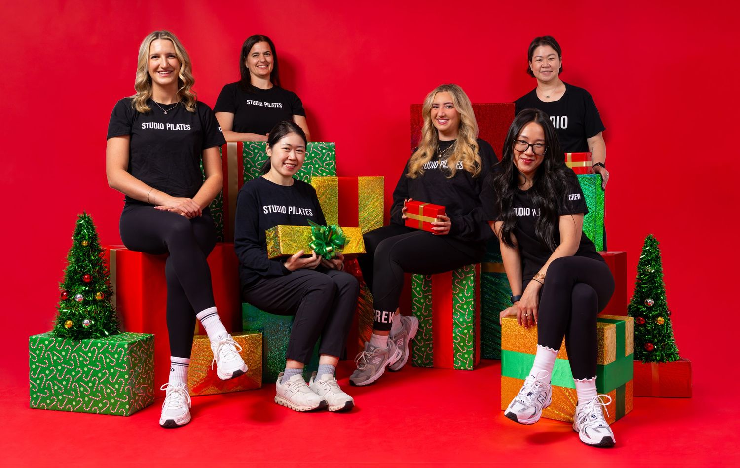 Six women in matching black outfits pose with colorful Christmas gifts and mini trees against a festive red background.