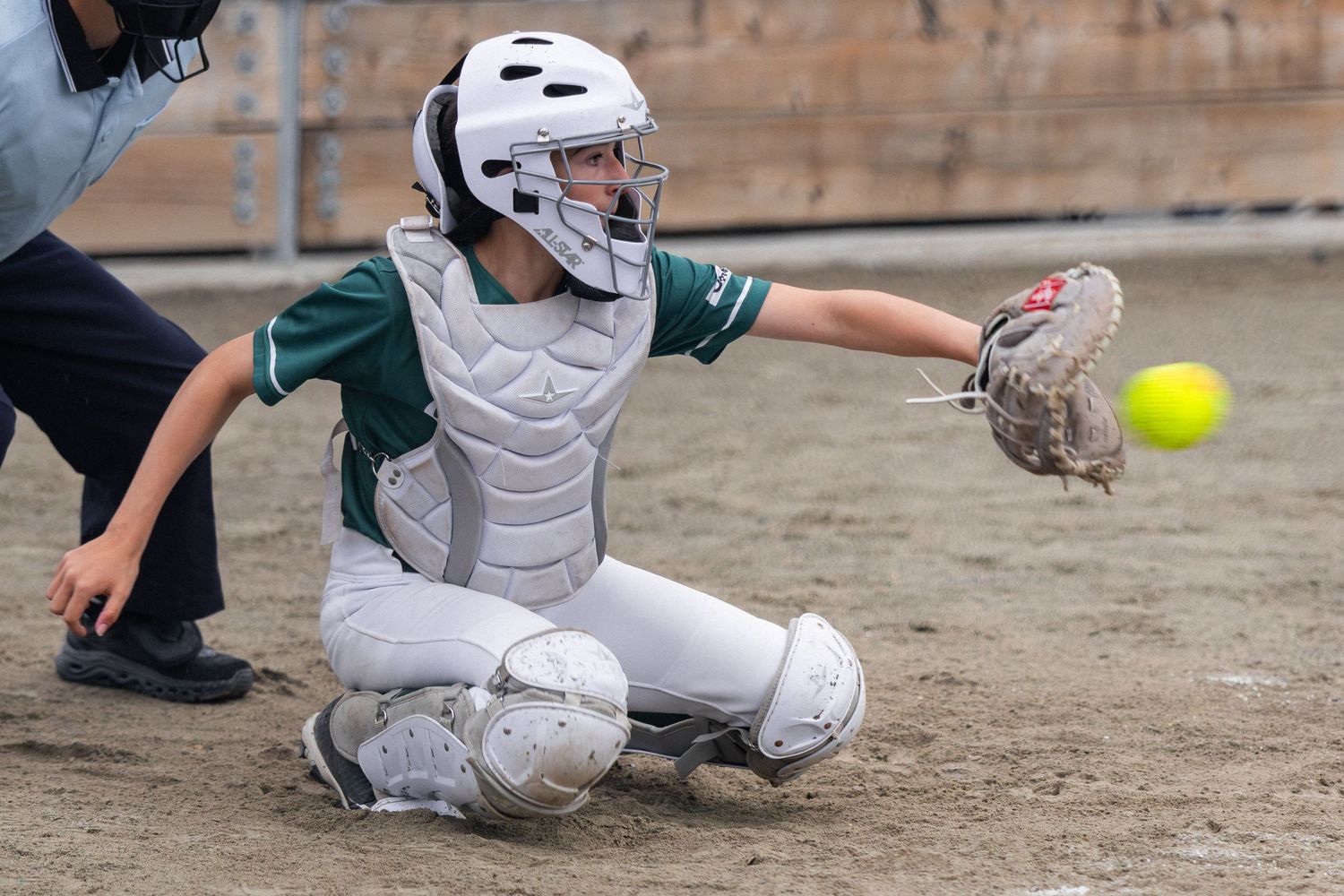Female softball catcher in green uniform crouching to catch a yellow ball during a game.