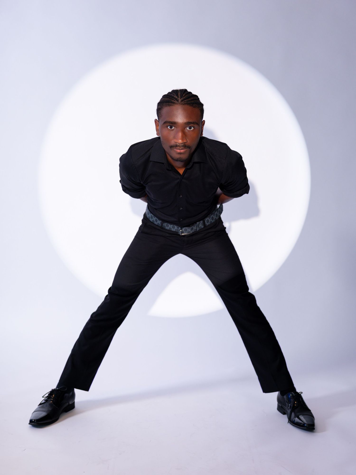 Young Black man in all-black outfit striking a wide-stance pose against a white circular light backdrop.
