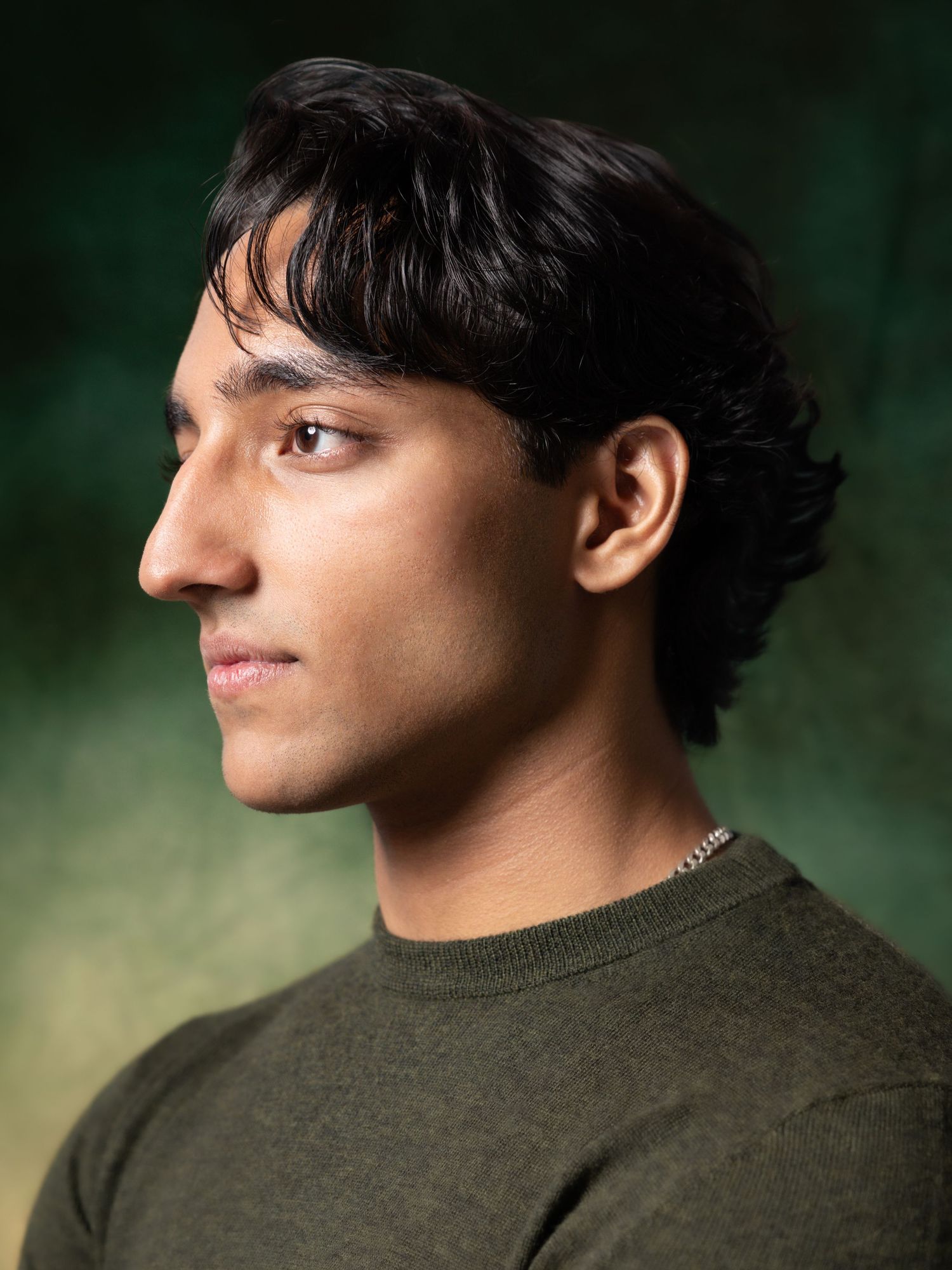 Young man in olive green sweater with silver chain necklace poses in profile against dark green background.