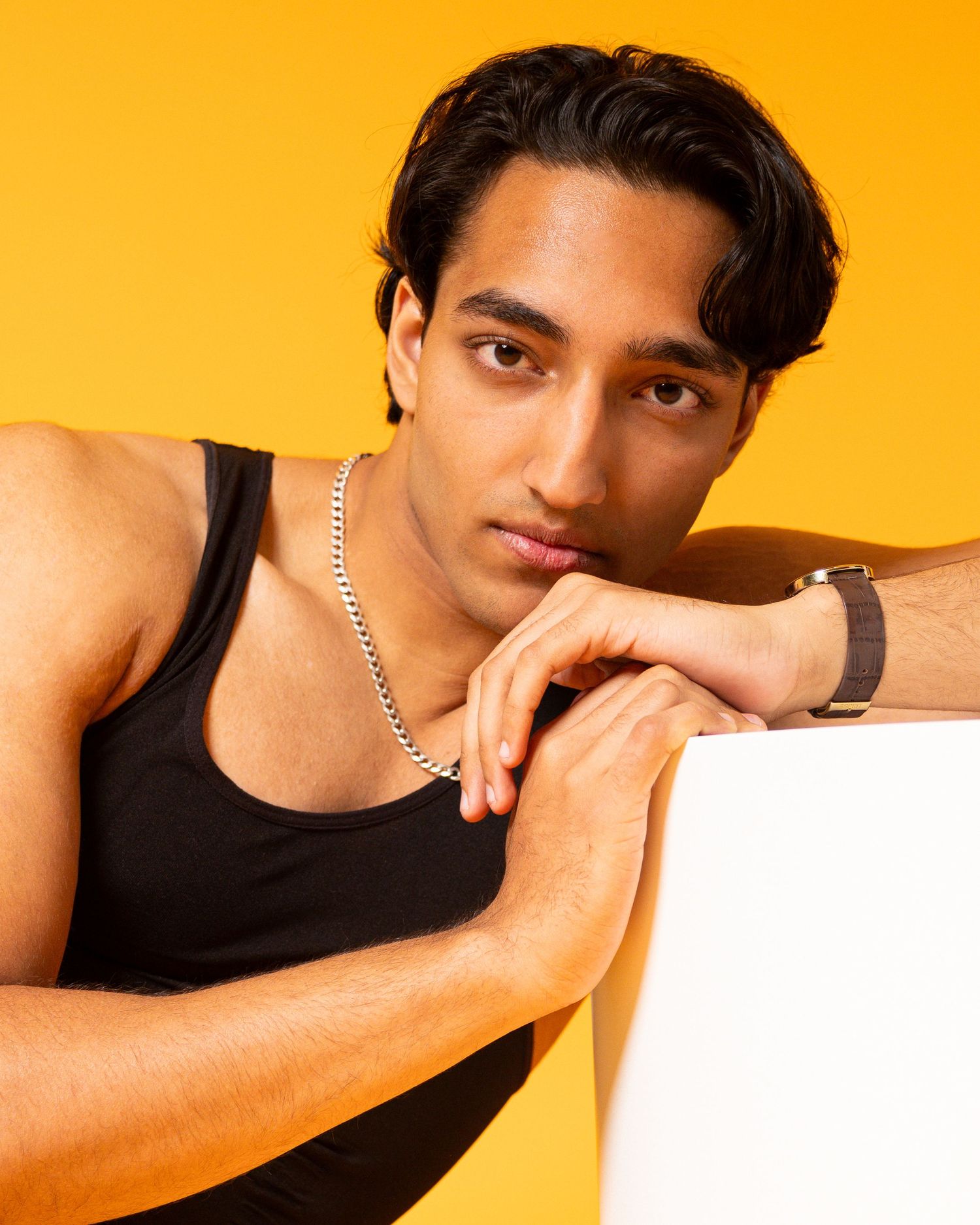Young man in black tank top and silver chain necklace poses against vibrant yellow background.