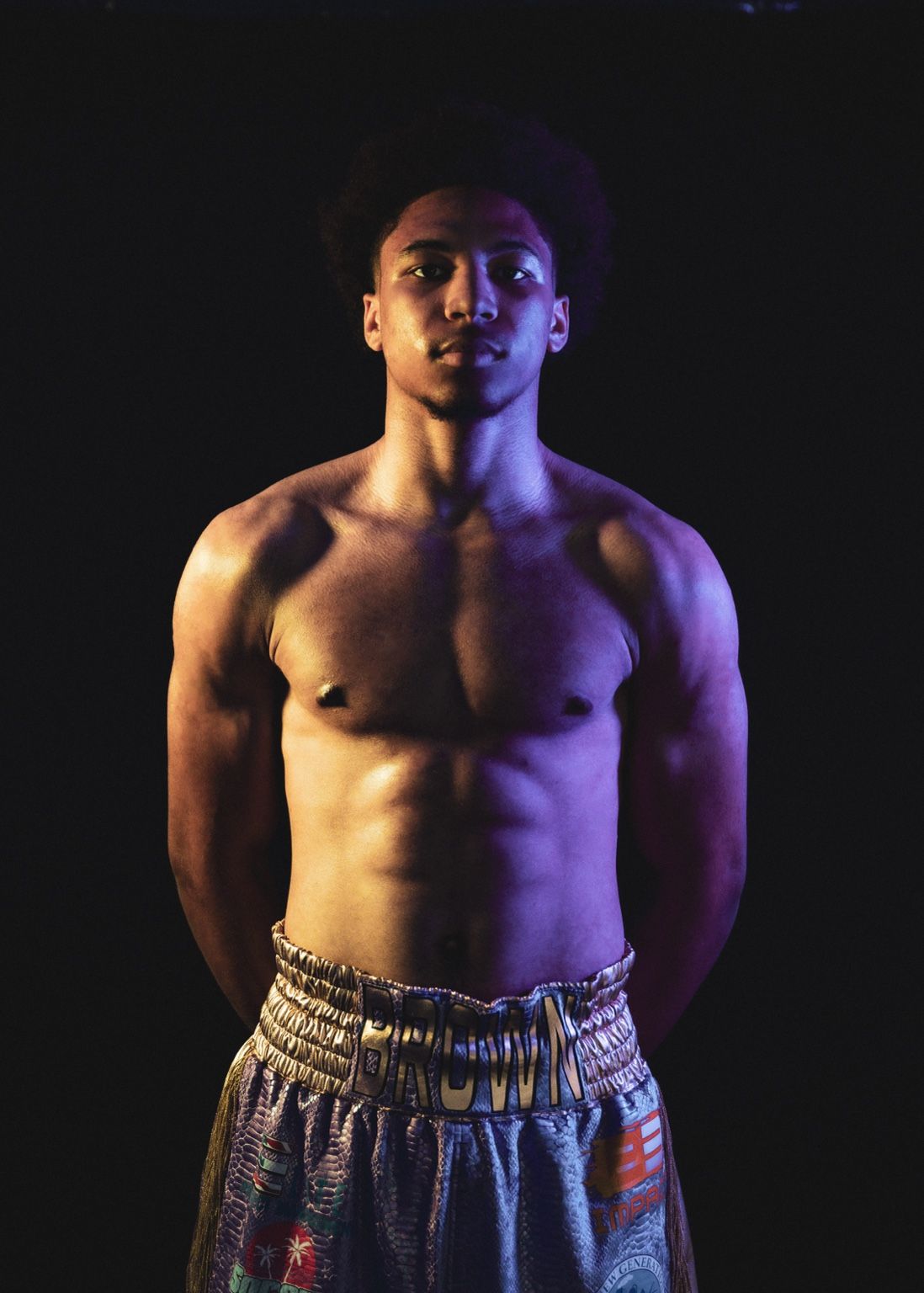 Shirtless muscular young man posing confidently against dark background with colorful patterned shorts.