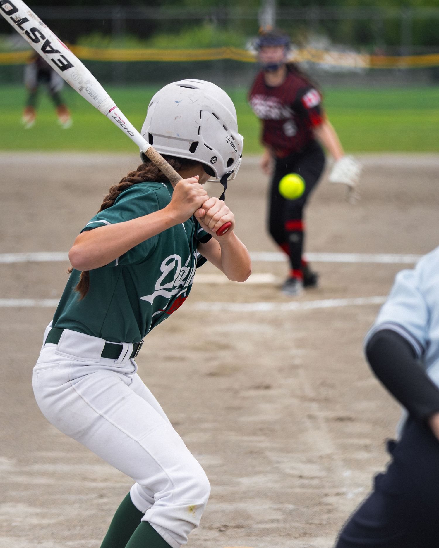 Young softball batter in green uniform swings at incoming yellow pitch during competitive youth softball game.