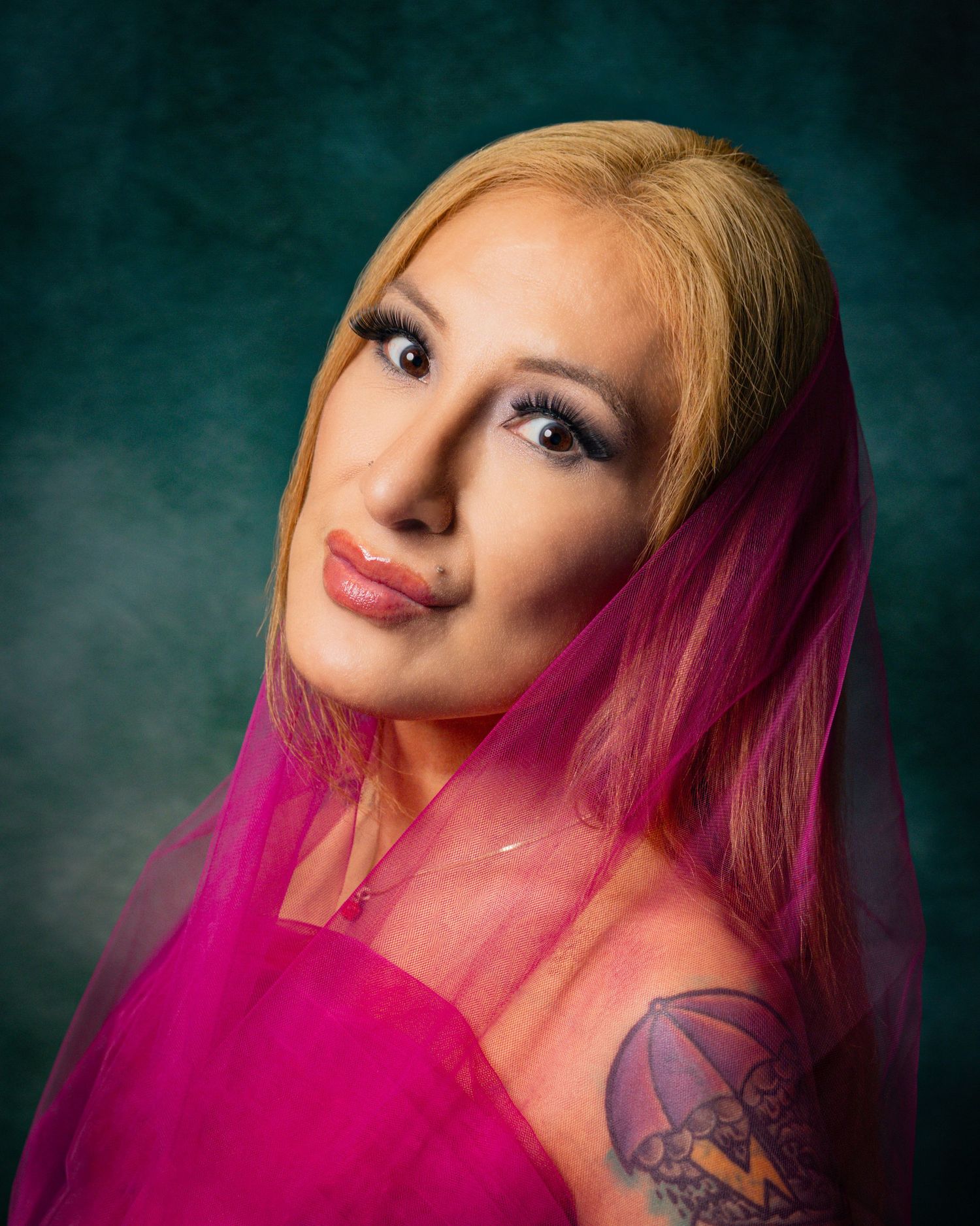 Woman with blonde and pink hair, blue eyes, bold makeup, and a tattoo, wearing a pink outfit against a teal backdrop.