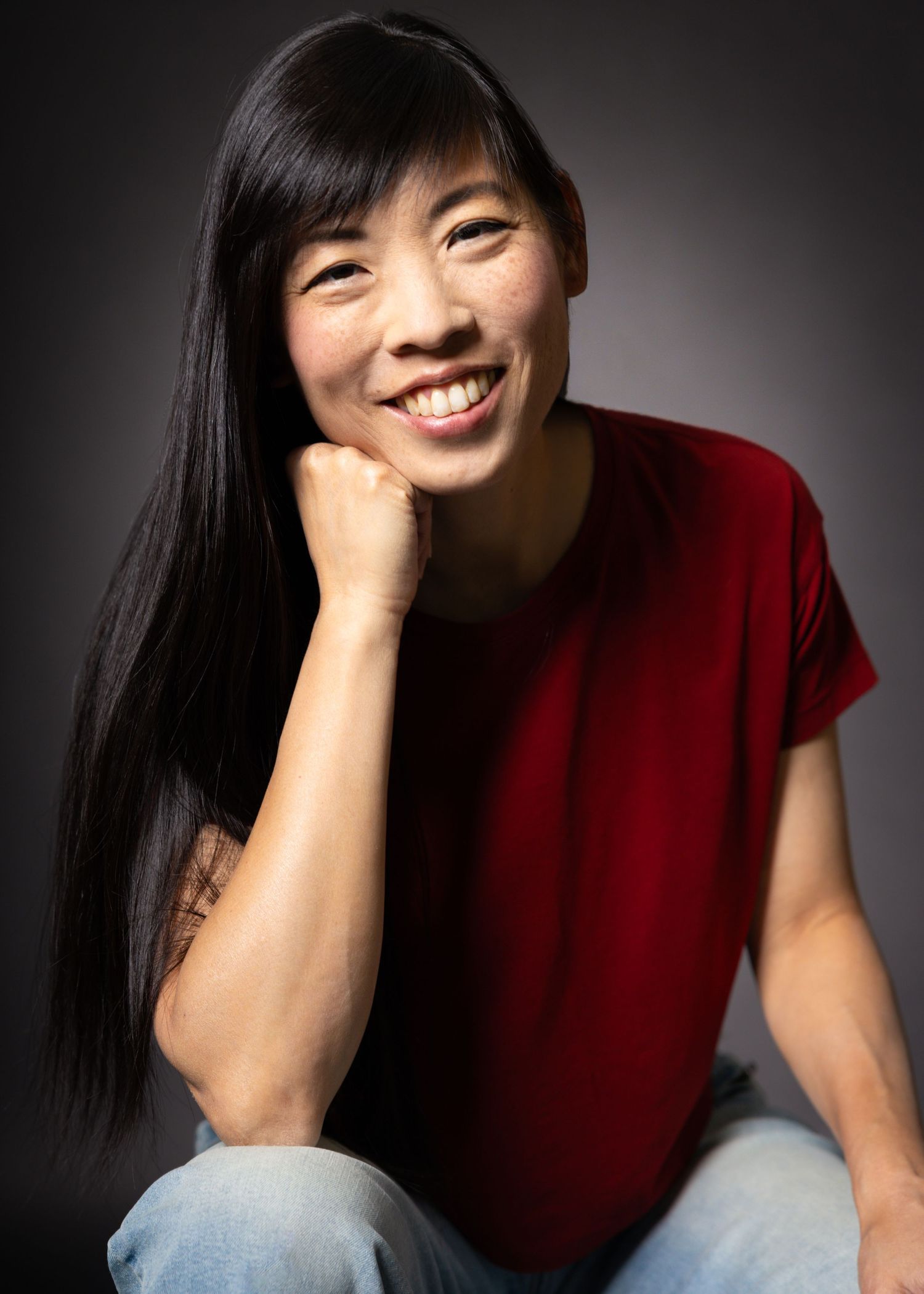 Smiling Asian woman in red t-shirt and jeans, resting chin on hand, posed against dark studio background.