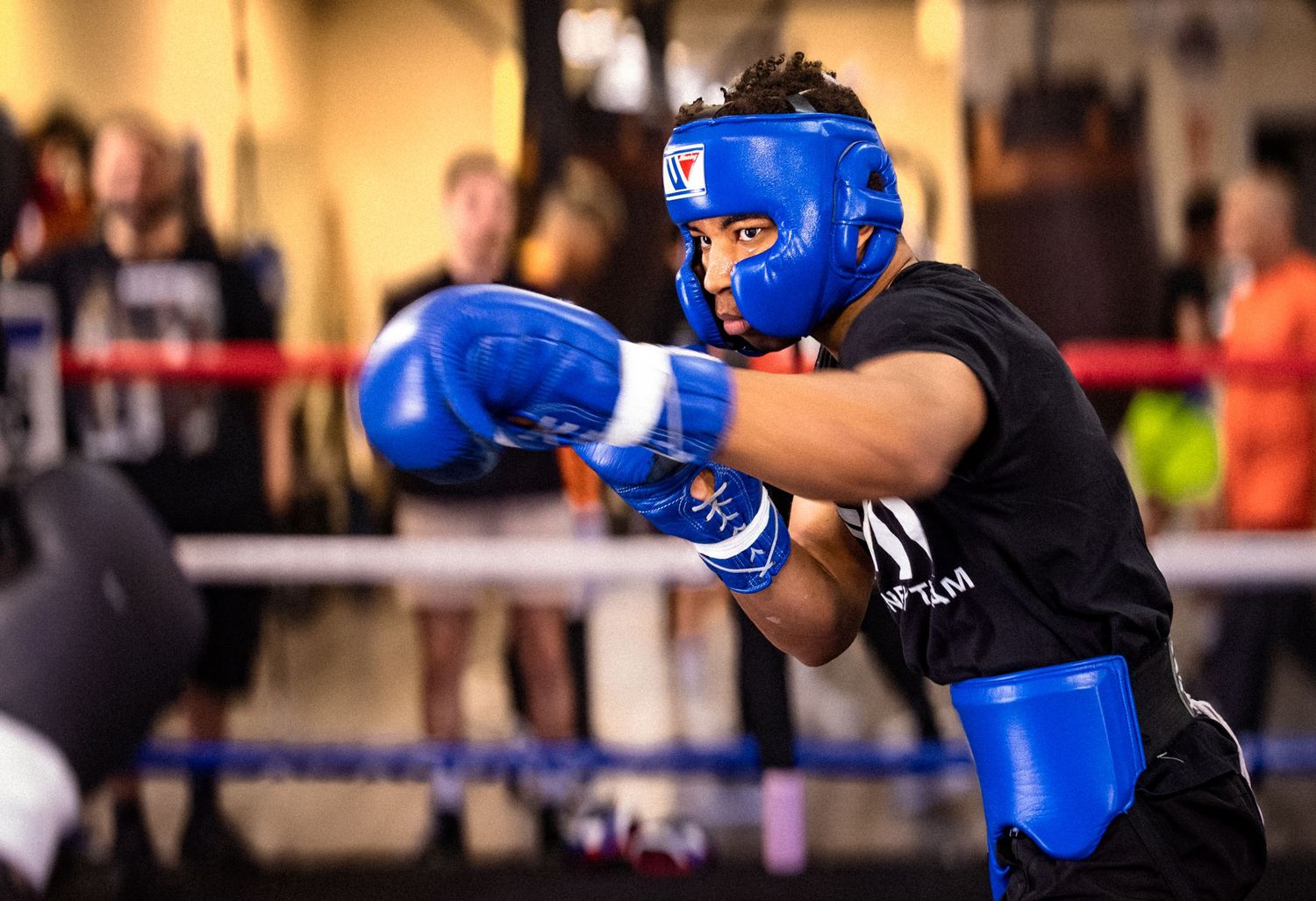 Young boxer in blue headgear and gloves training in boxing ring during sparring session.