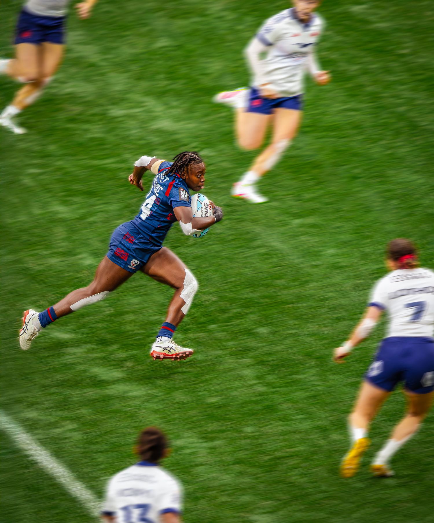 Female rugby player in blue jersey sprinting with ball during competitive match on green field.