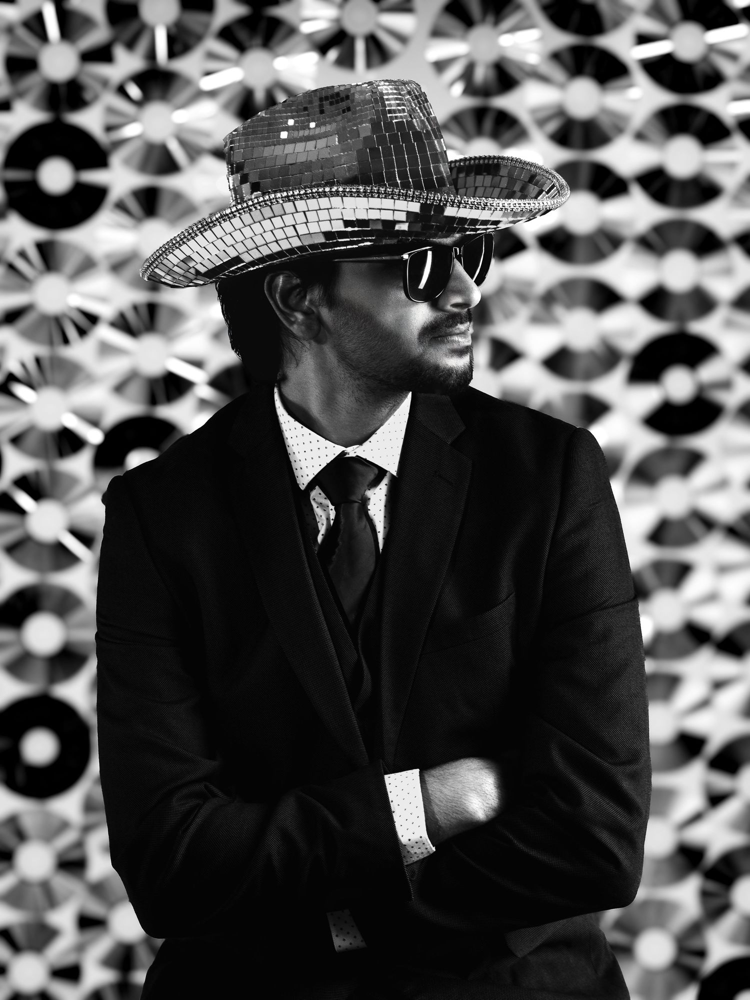 Black and white portrait of a stylish man in a suit, sunglasses, and sequined wide-brim hat with arms crossed.