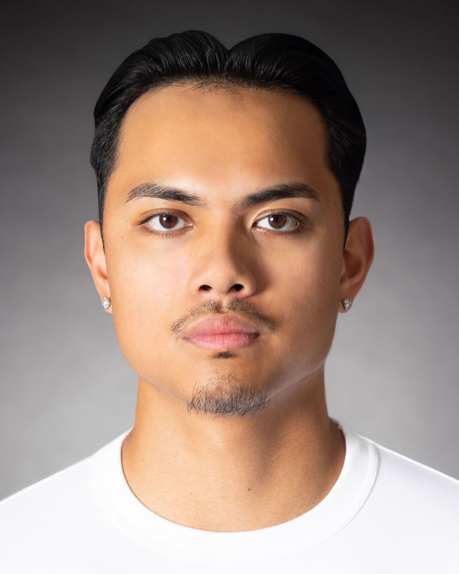 Young man with dark hair, small earrings, and a white t-shirt poses for a professional headshot photo.