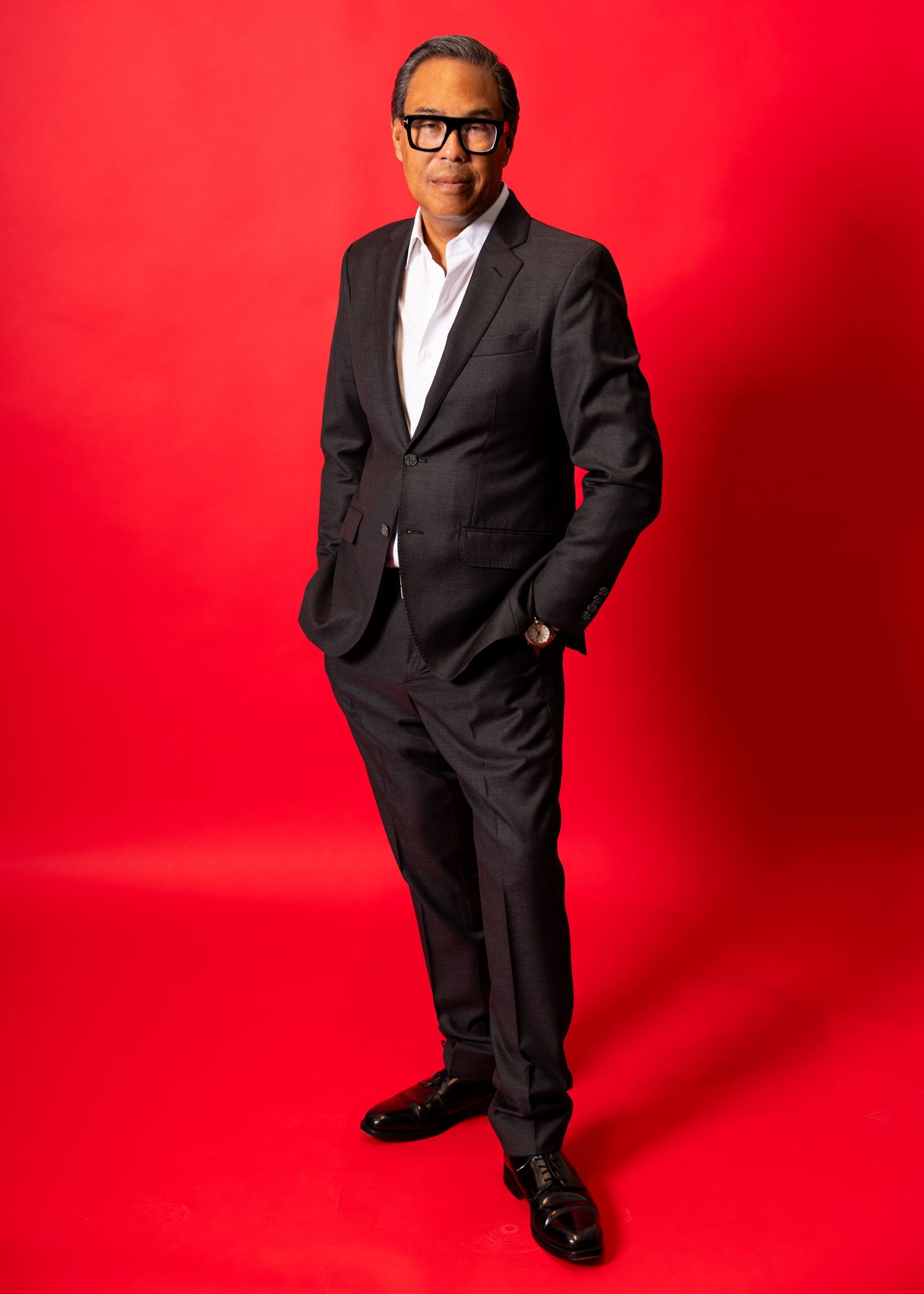 Man in dark suit and glasses poses confidently against a bold red background in a professional portrait photo.
