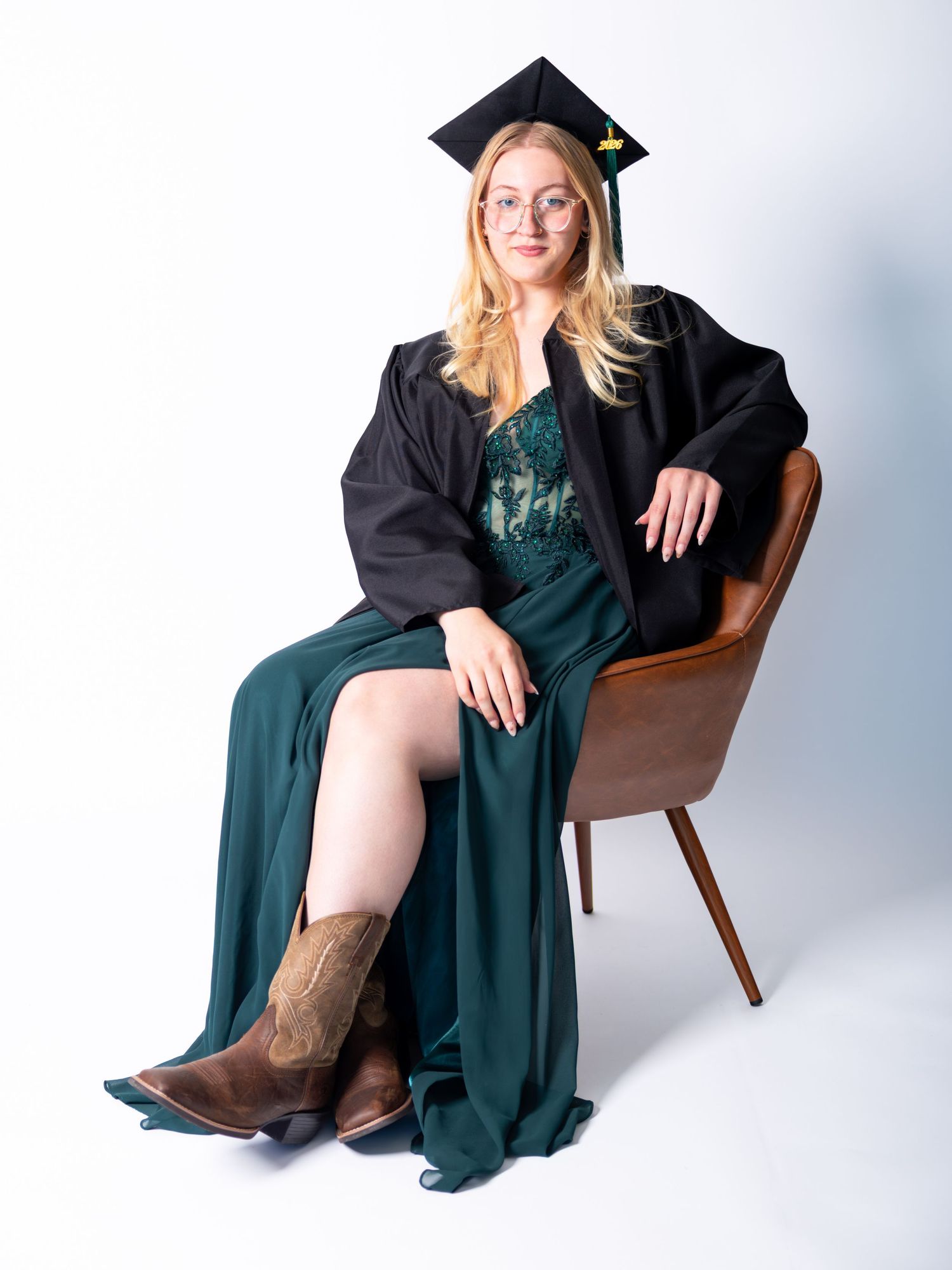 Smiling blonde graduate in black cap and gown over green dress, seated in brown chair, wearing glasses and cowboy boots.