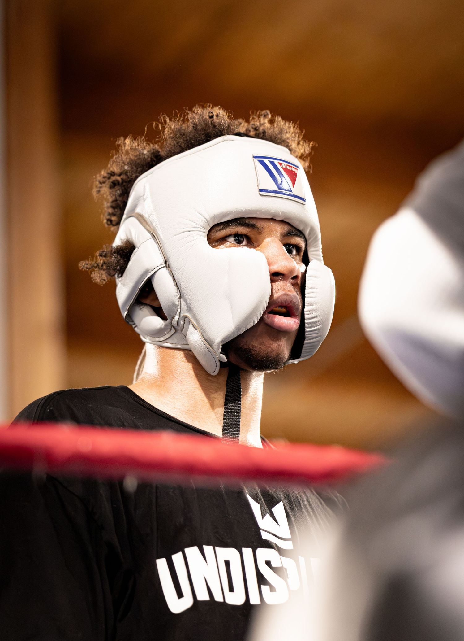 A boxer wearing a white Winning headgear and black Undisputed shirt trains in a boxing ring.