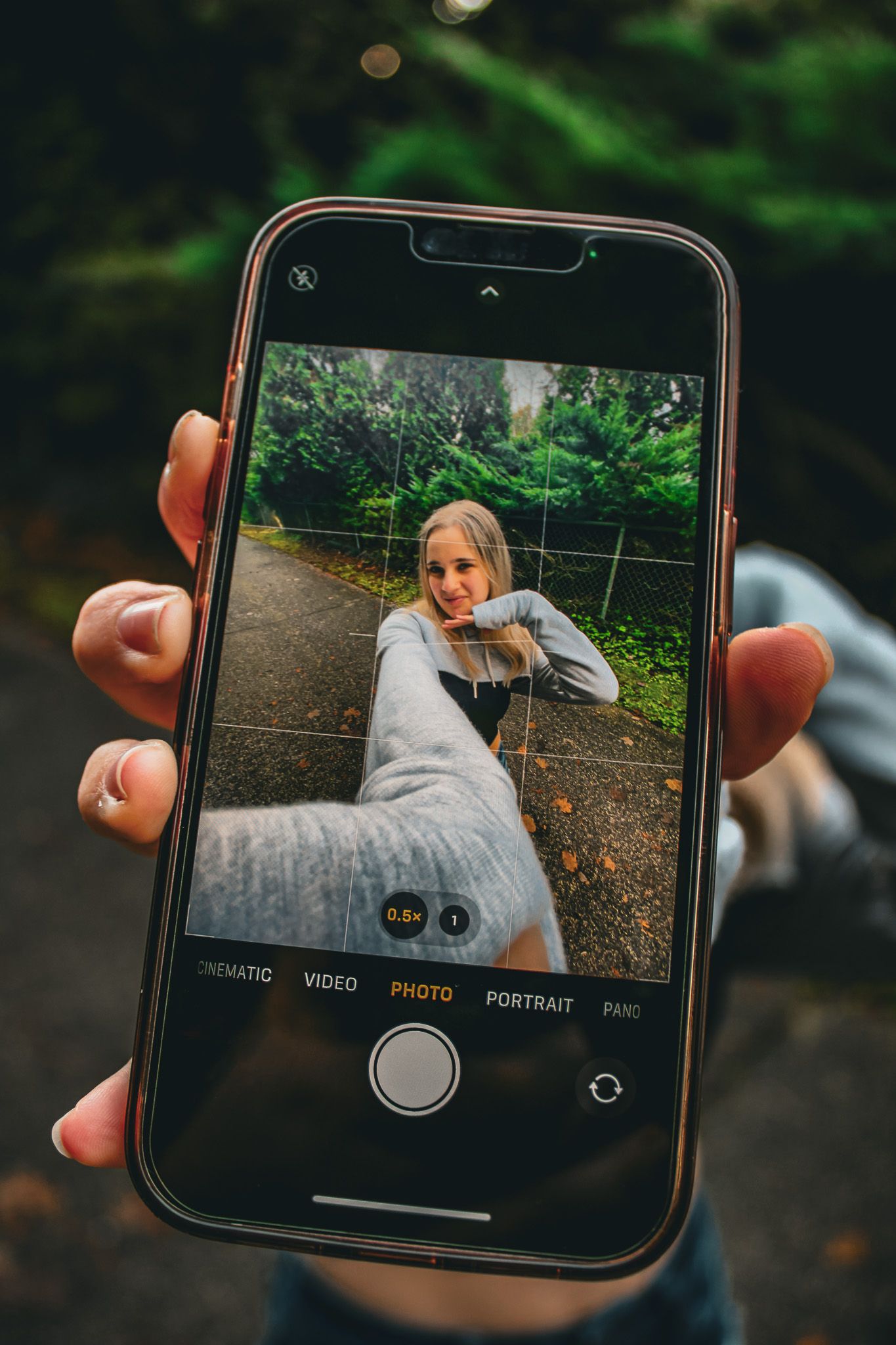 Person holding an iPhone photographing a smiling blonde woman on a forest path in portrait mode.