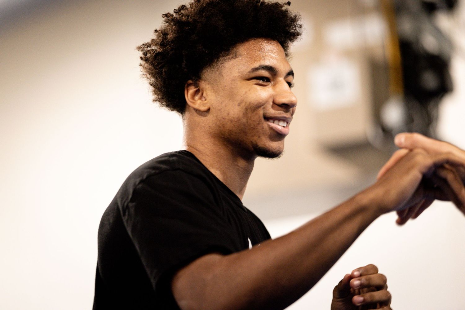 Young man in black t-shirt smiling and gesturing in a bright indoor setting.