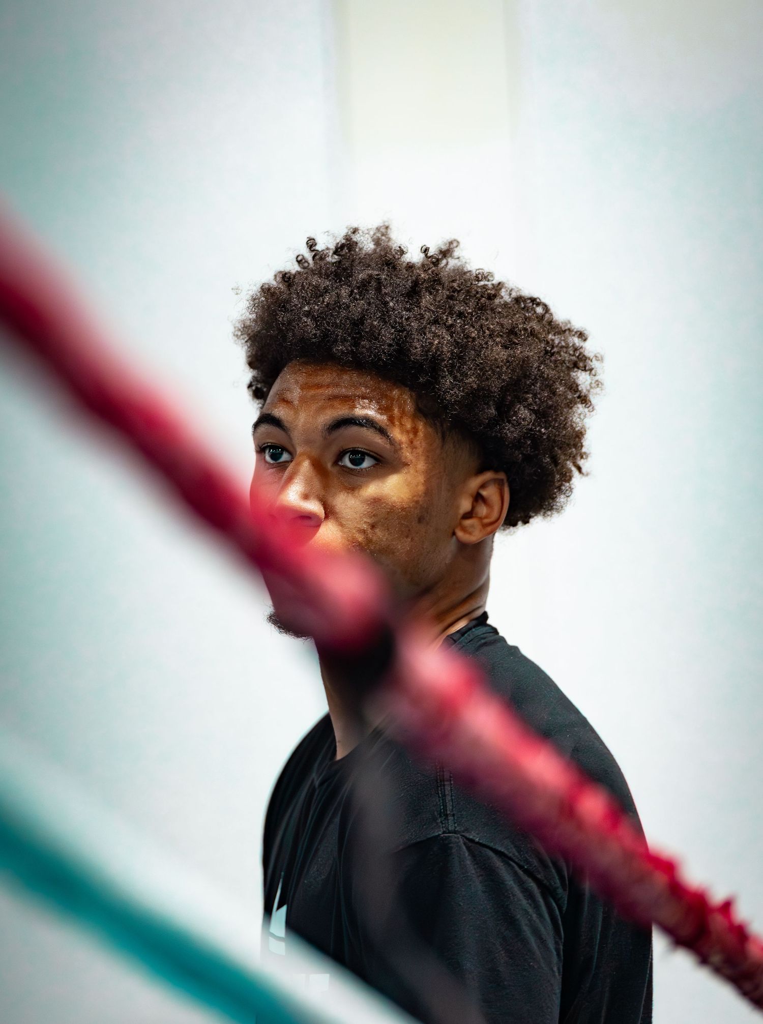 Young boxer with curly hair and bruised face standing behind red ring ropes in a gym.