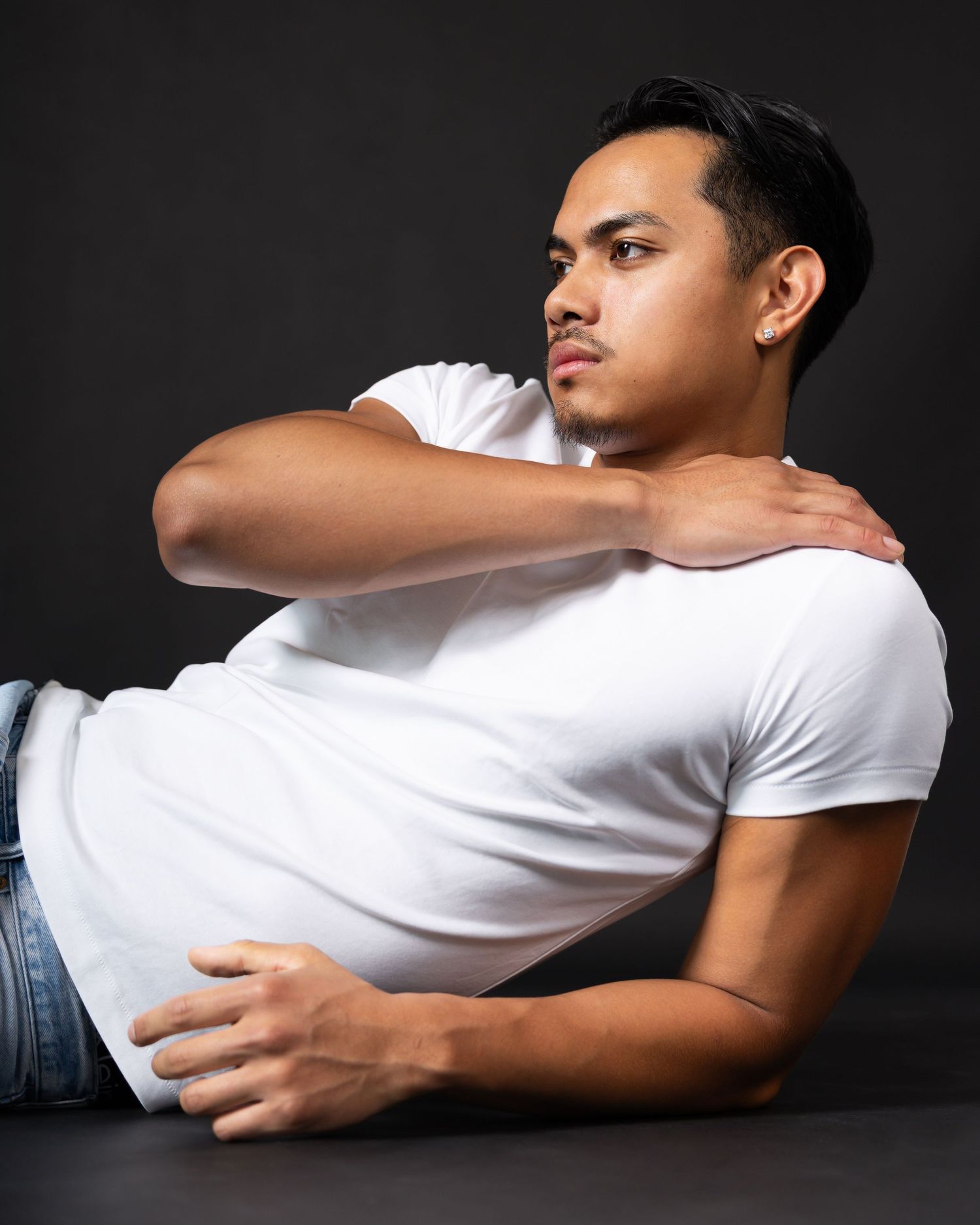 Young man in white t-shirt and jeans poses lying down against dark background, looking sideways with hand on shoulder.