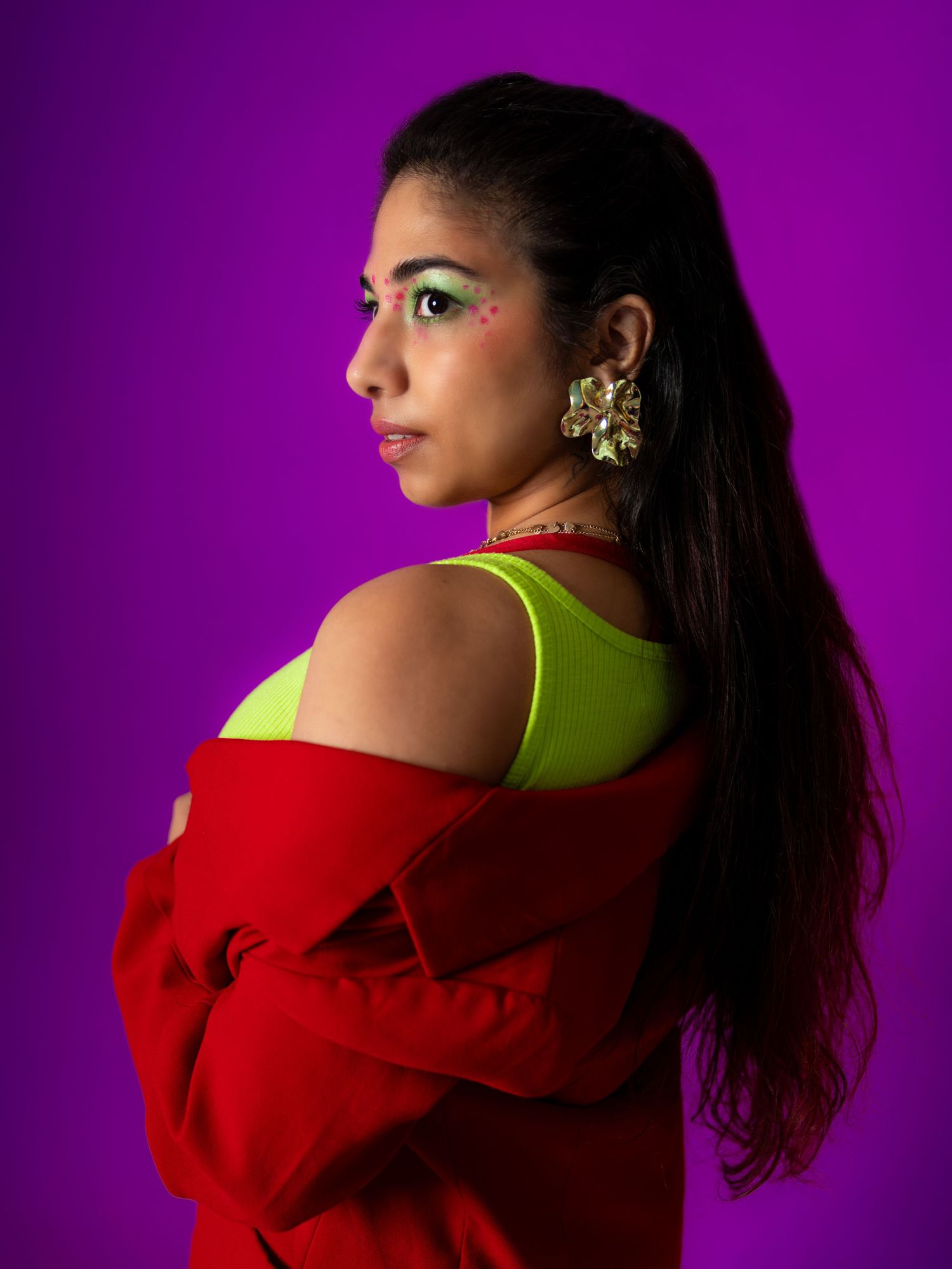 Woman in red jacket and neon yellow top with bold green eye makeup and gold earrings posing against purple background.