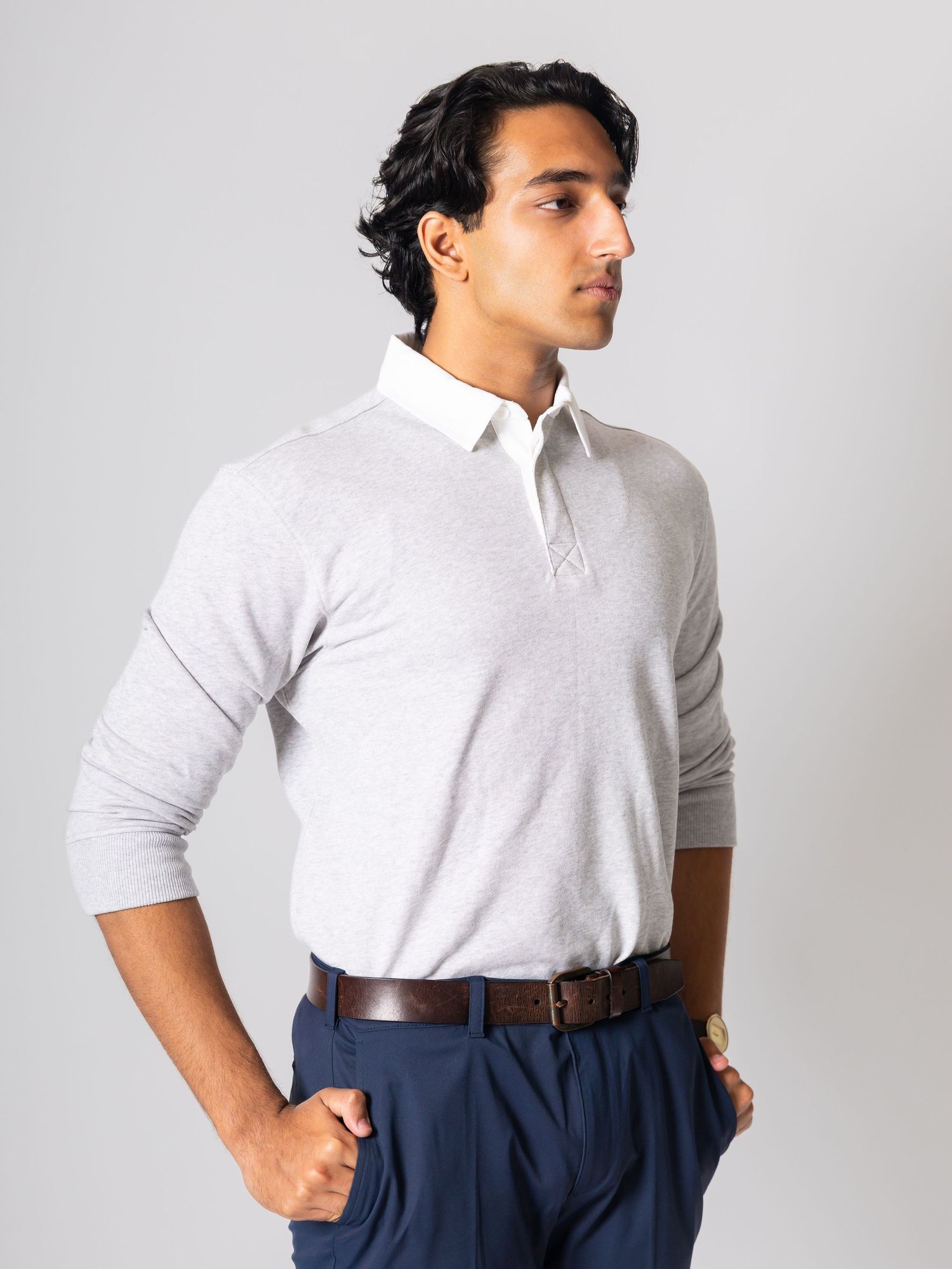 Man wearing a gray long-sleeve polo shirt with white collar, navy trousers, and brown belt against a gray background.