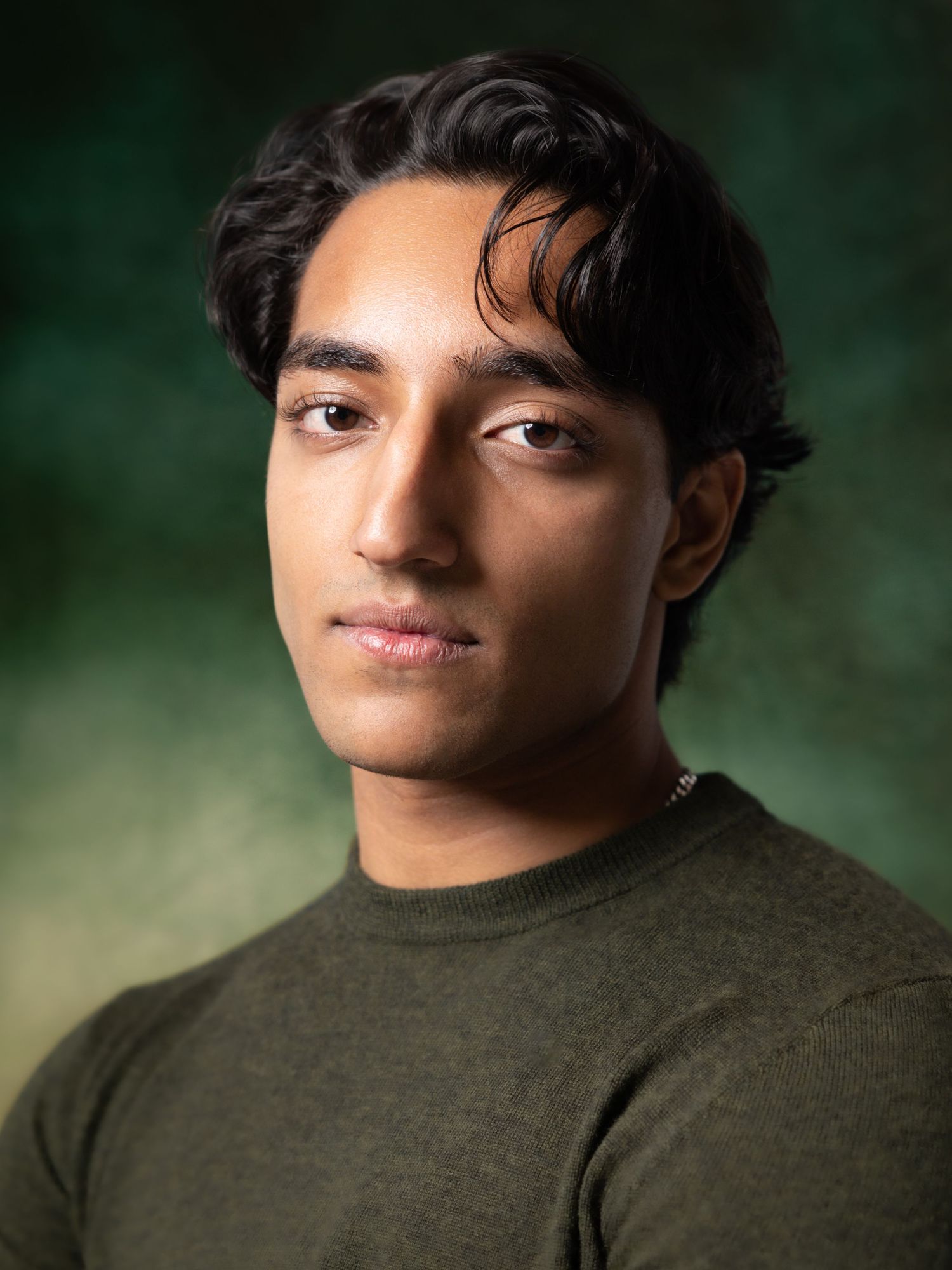 Young man with dark hair wearing an olive green sweater poses for a professional headshot portrait.