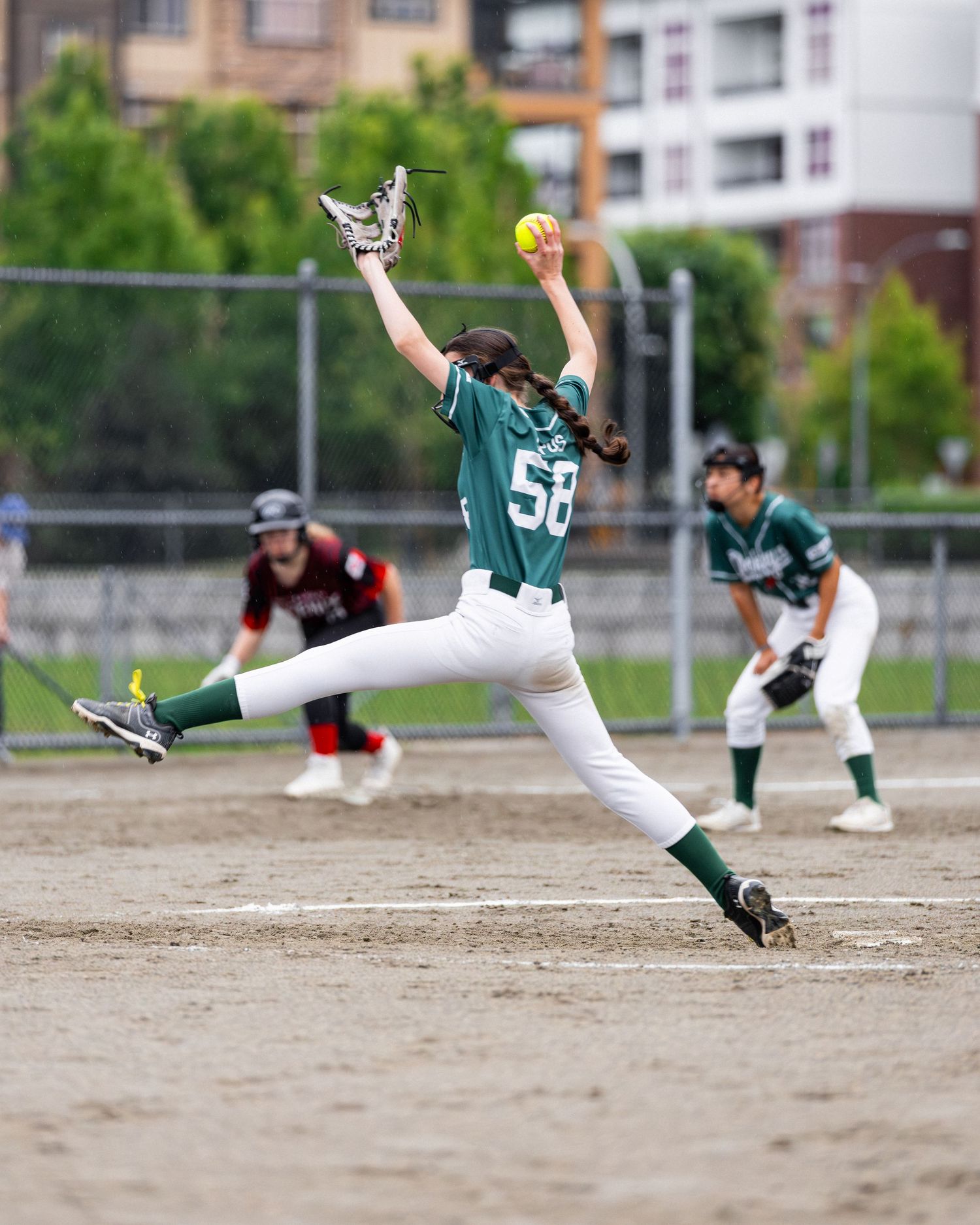 Softball pitcher in green jersey #58 winds up to throw during a game on a dirt field with teammates visible in background.