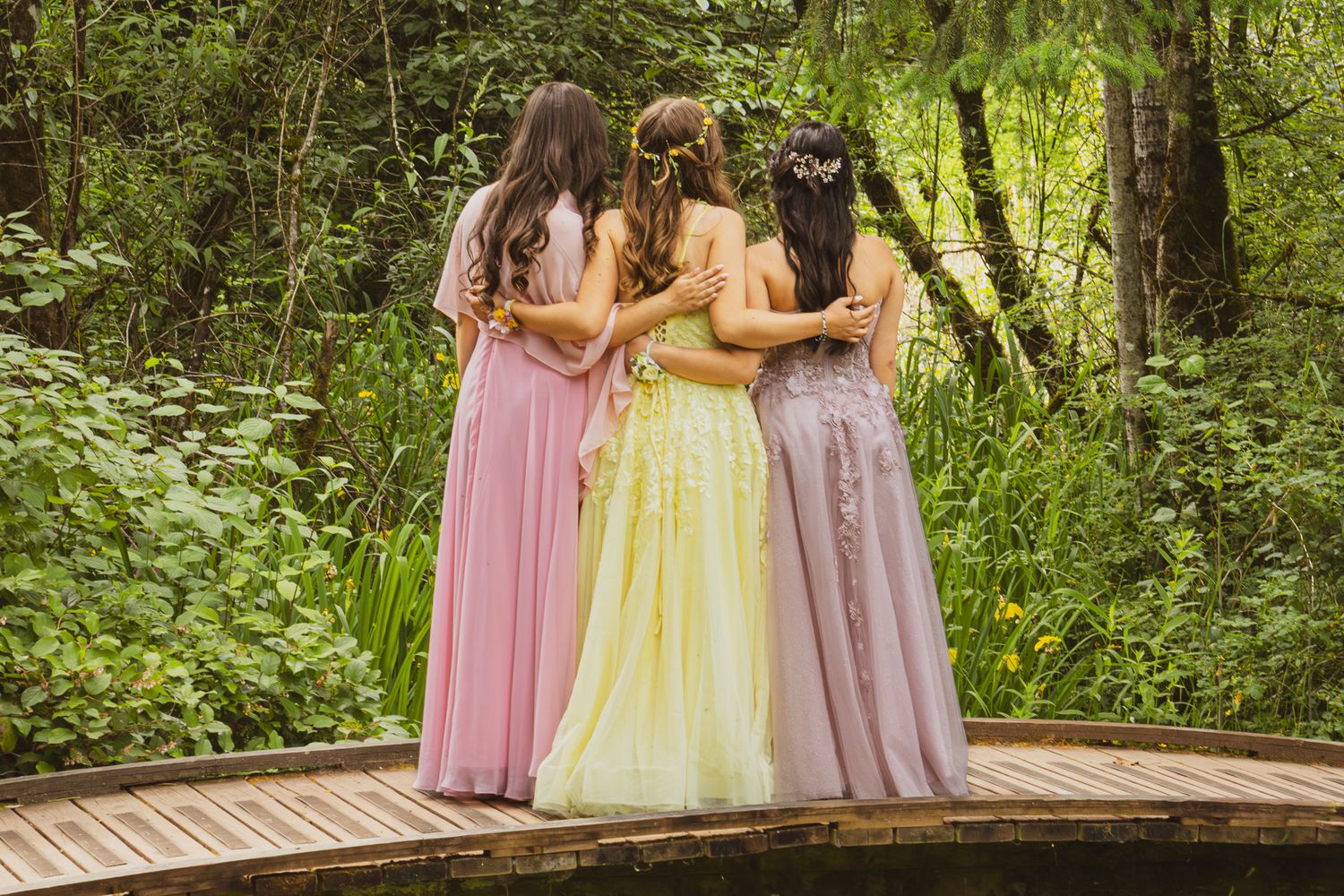 Four bridesmaids in pastel gowns stand embracing on a wooden bridge surrounded by lush greenery.