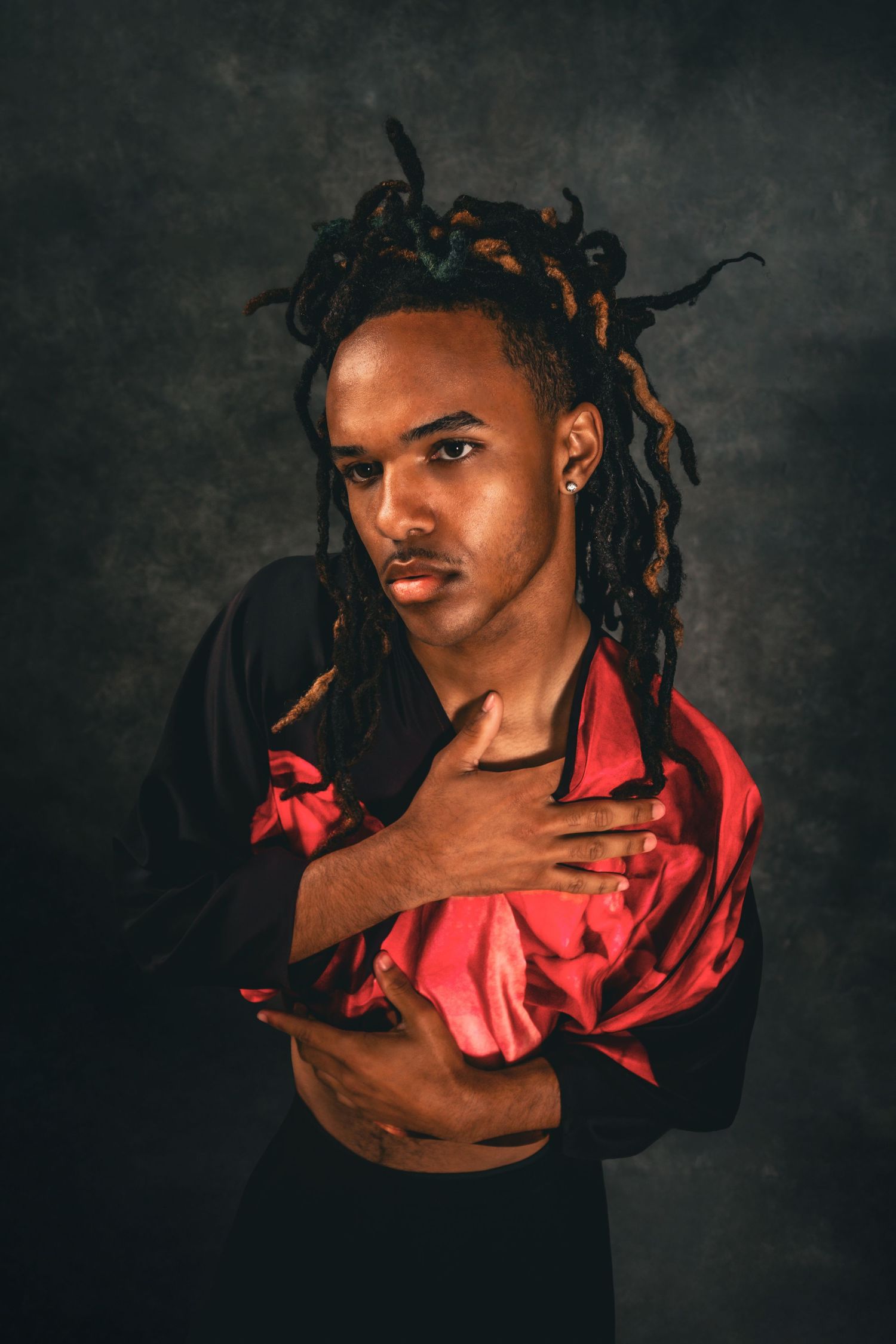 Young Black man with colorful dreadlocks poses in black and red satin shirt, crossing arms over chest in dramatic studio portrait.