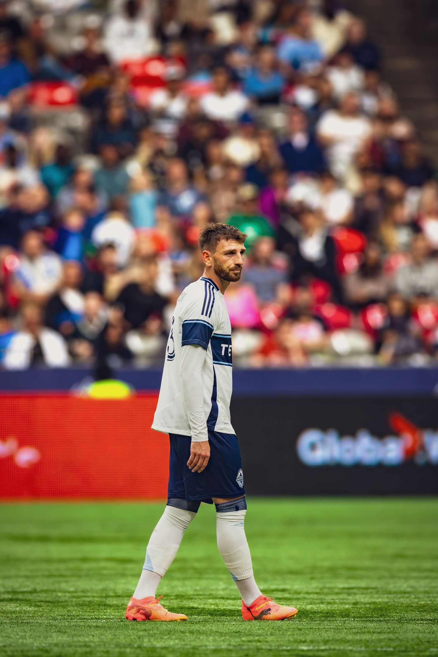 Male soccer player in blue and white uniform walks on green field during a professional match with crowd in background.