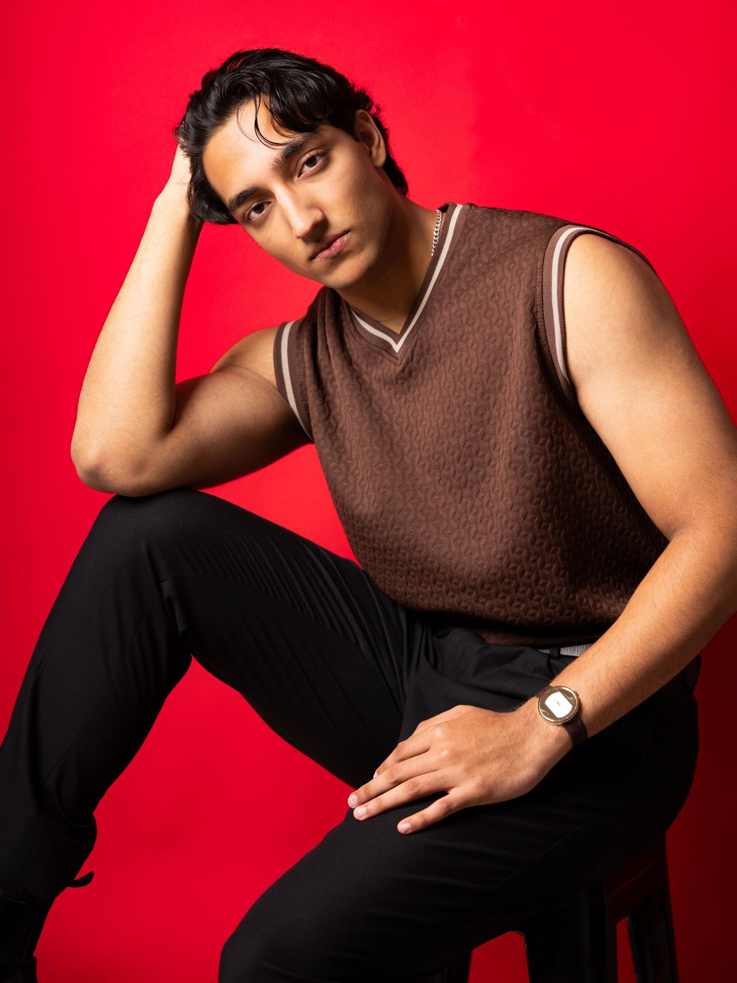 Young man in brown sleeveless top and black pants poses on stool against red background.
