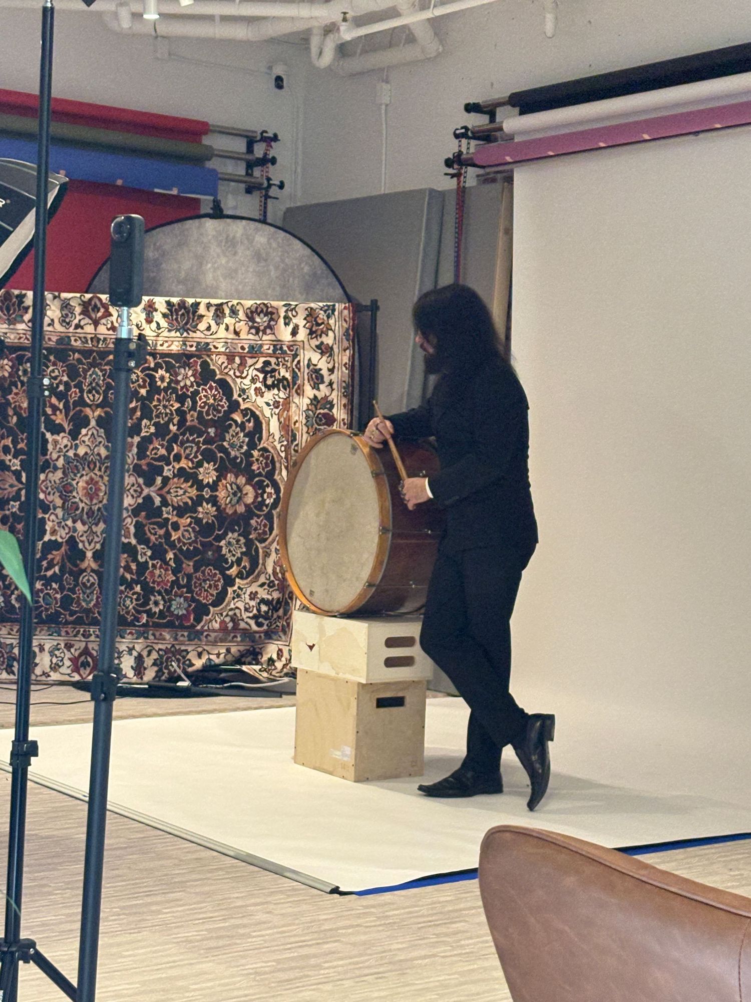 Musician in black outfit playing a large traditional drum on a wooden stand in a photography studio.
