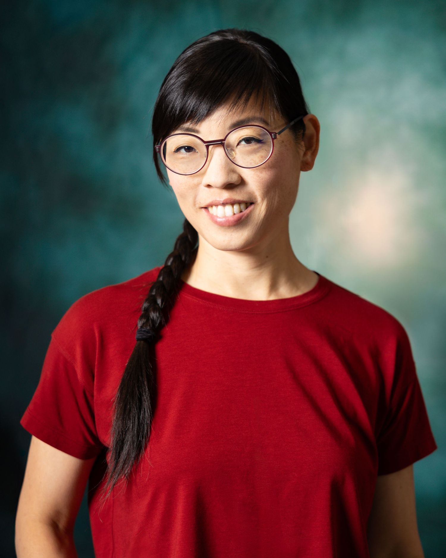Smiling Asian woman with braided hair and round glasses wearing a red t-shirt against a teal background.