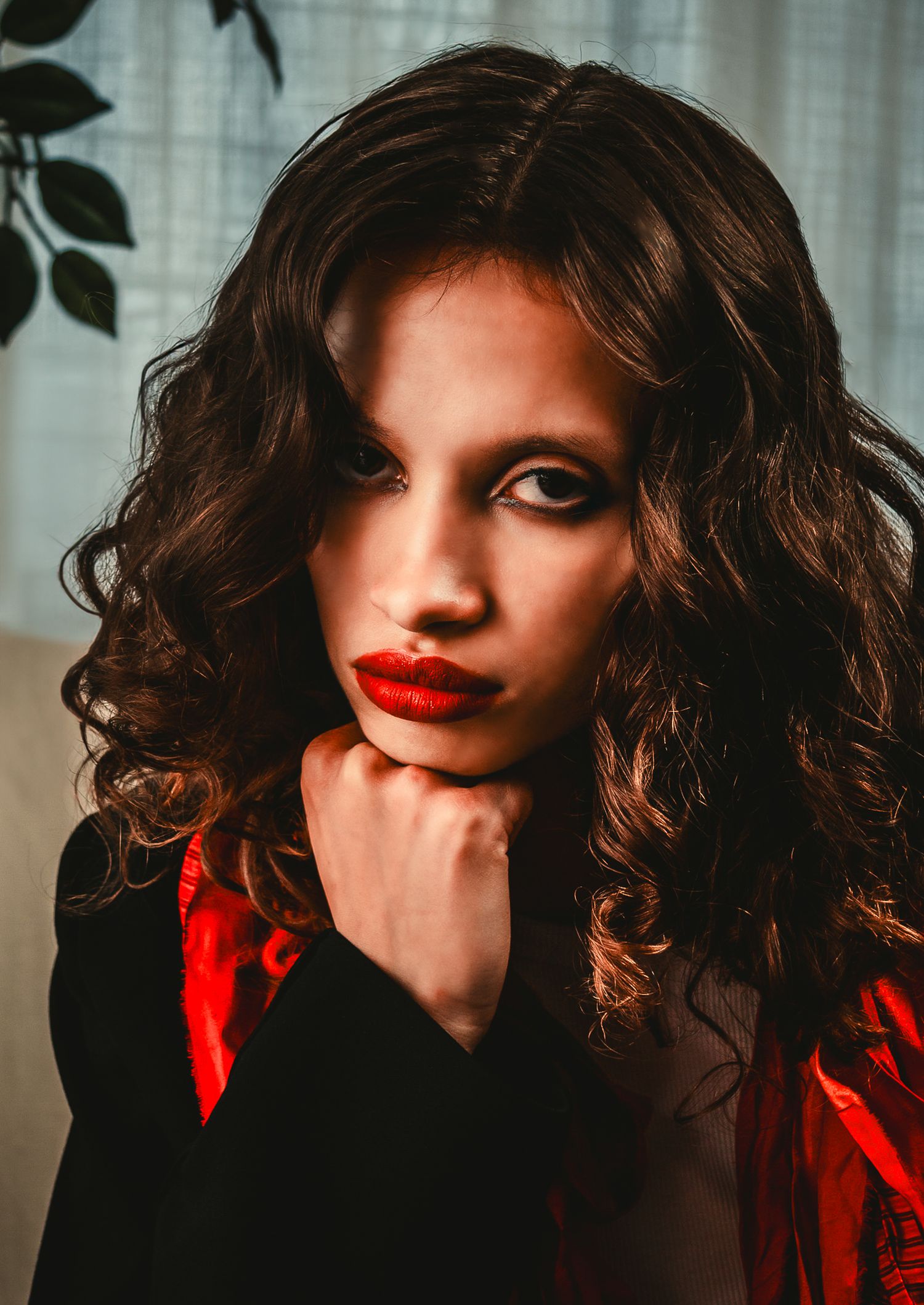 Young woman with curly dark hair and red lips resting chin on hand in dramatic portrait.