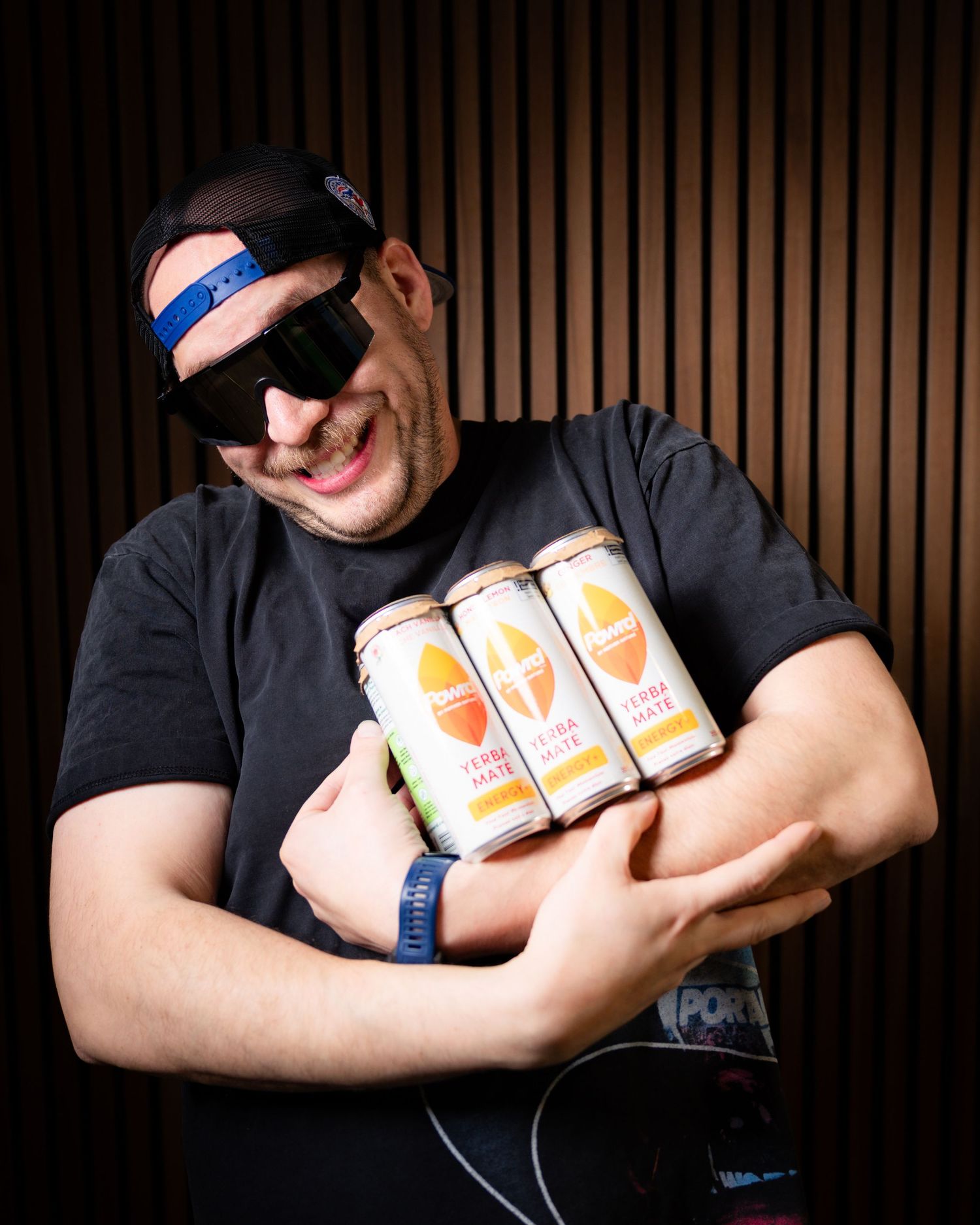 Man in sunglasses and black t-shirt hugging four cans of craft beer, smiling indoors.