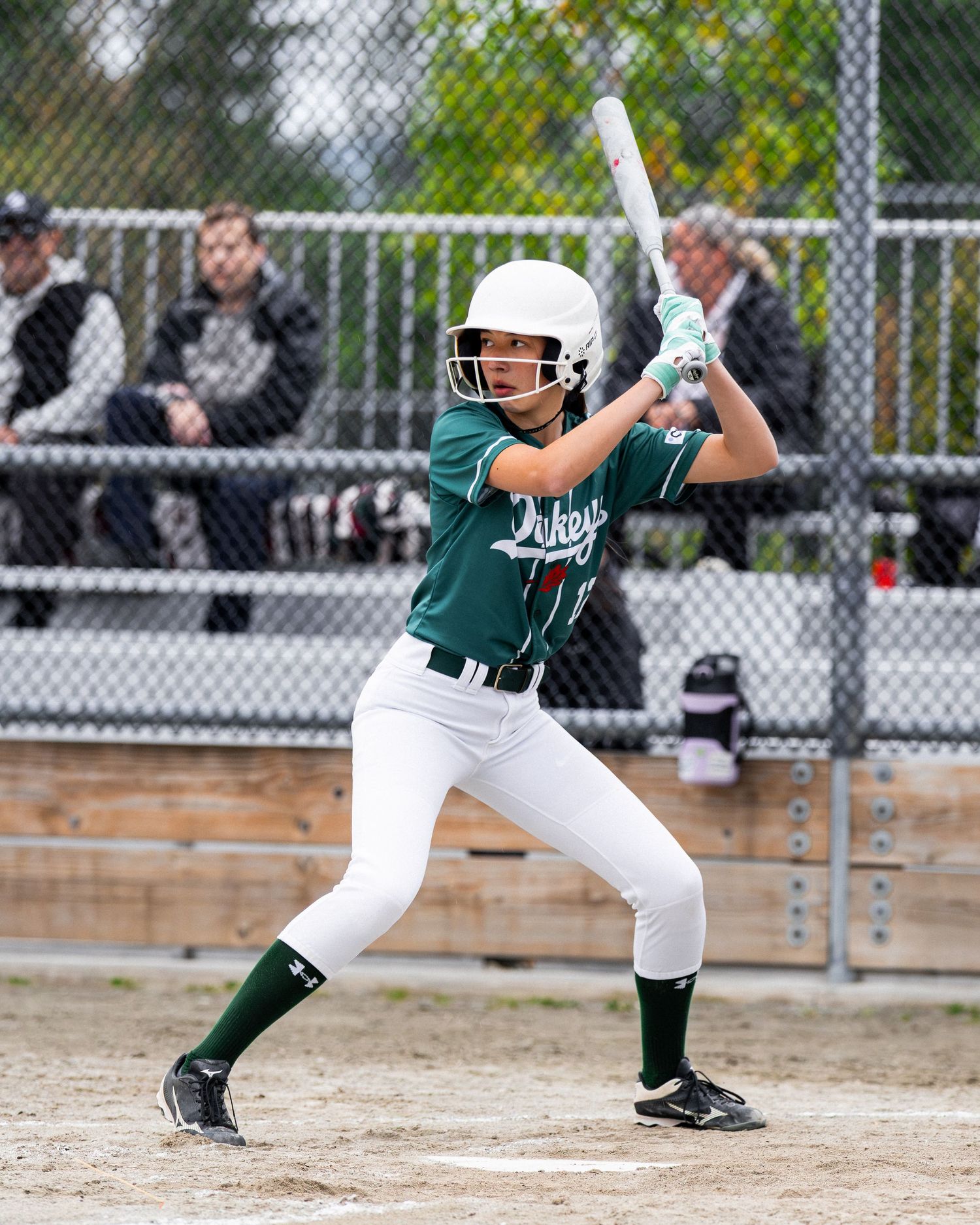 Female softball player in green uniform swings bat during a game, wearing helmet and cleats on an outdoor field.