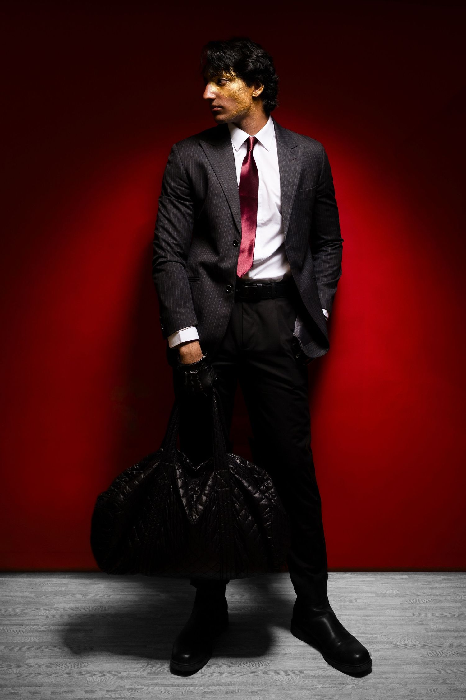 A stylish man in a dark suit and red tie holds a guitar case against a red wall.