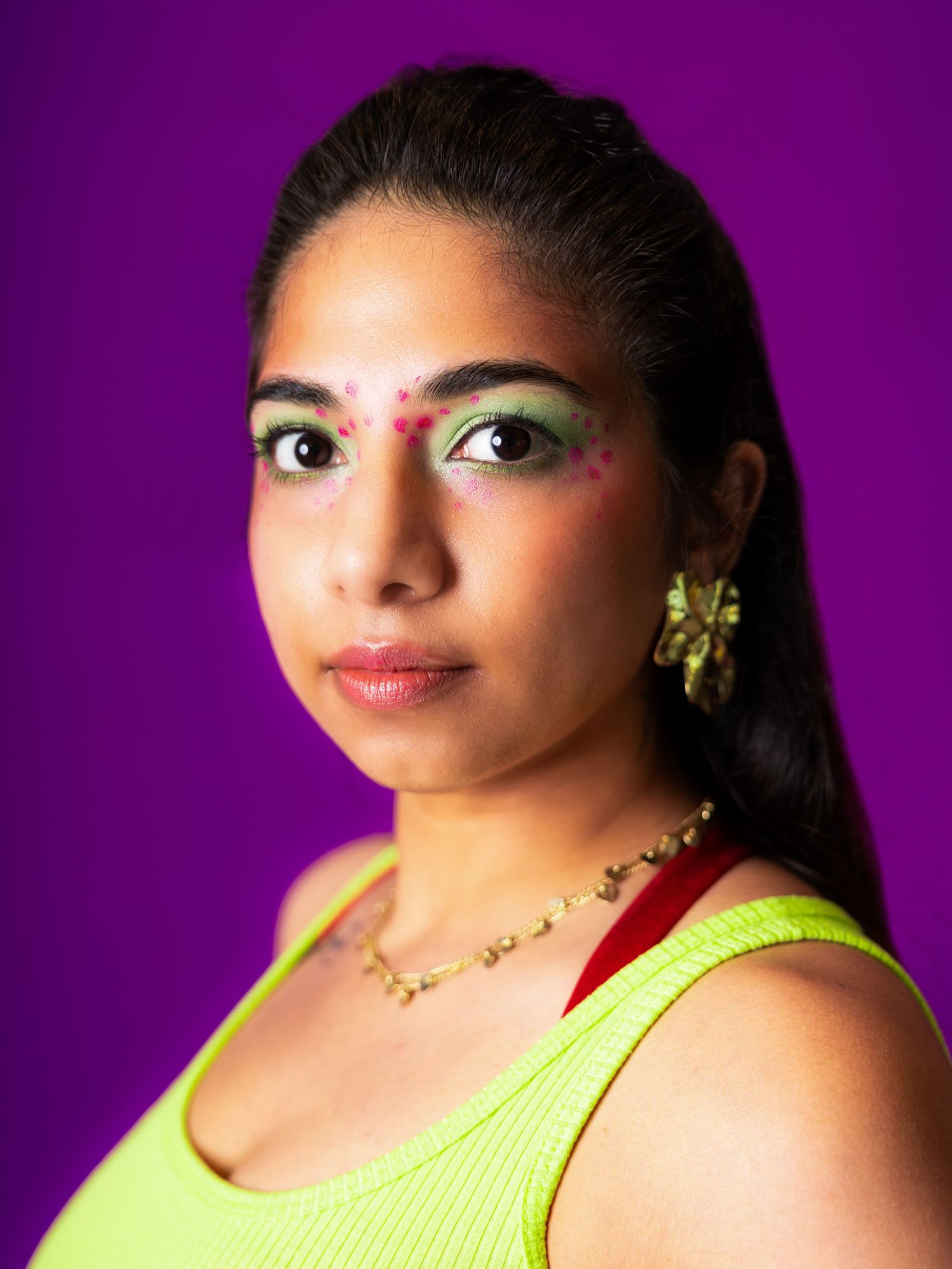 Woman with colorful eye makeup, bindi, gold jewelry, and neon yellow top poses against purple background.
