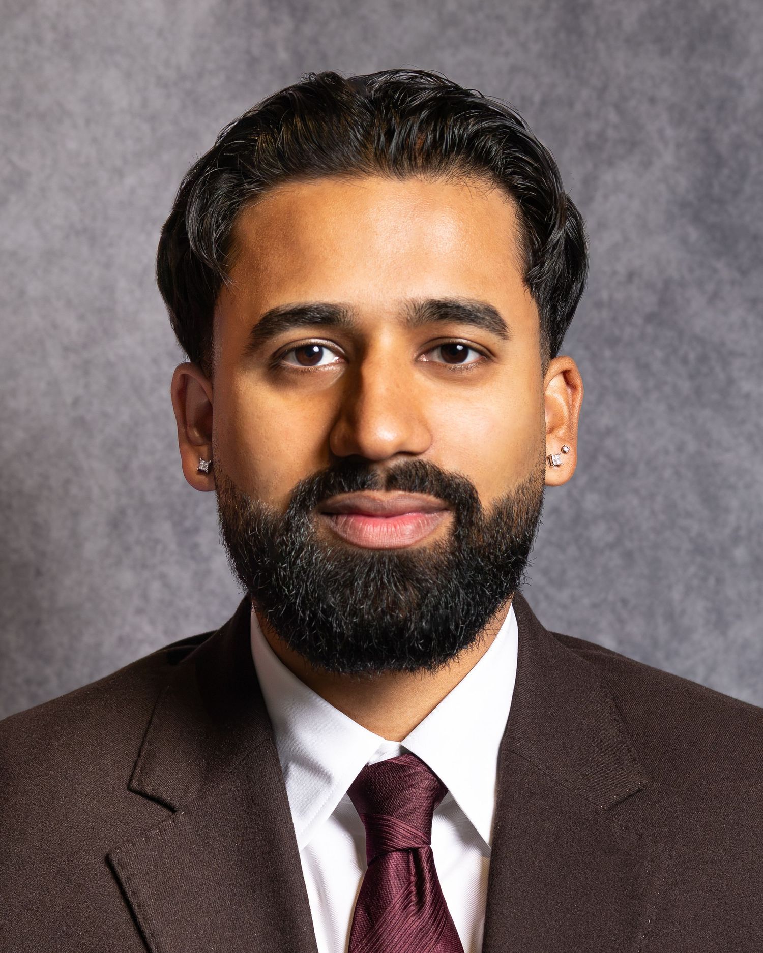 Professional headshot of a bearded man in a brown suit and burgundy tie with small earrings.