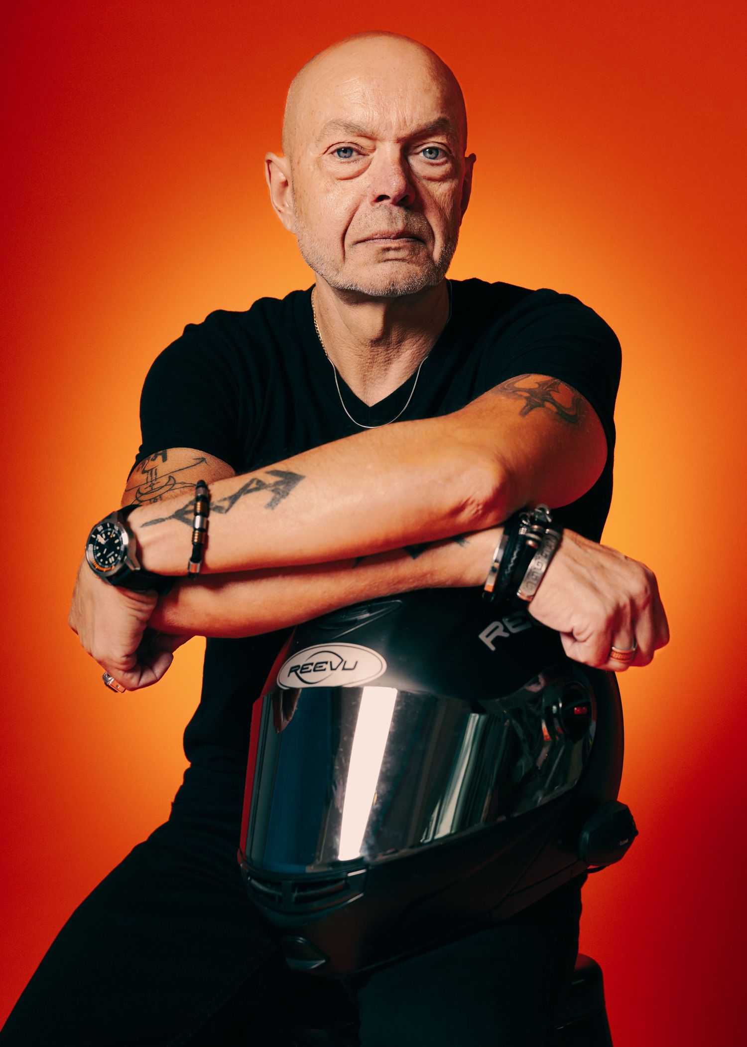 Bald tattooed man in black t-shirt poses with motorcycle helmet against vibrant orange-red background.