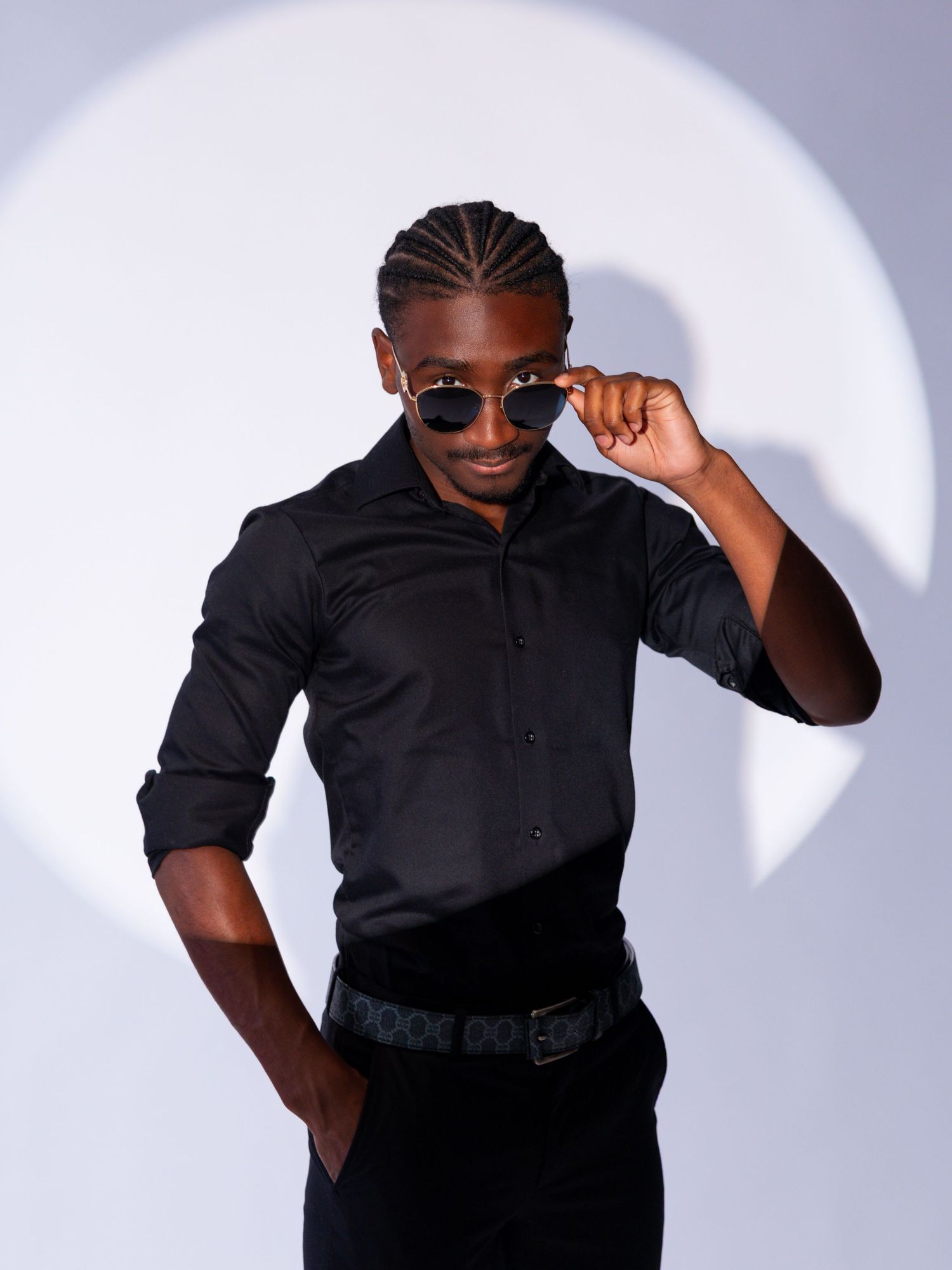 A stylish young man in a black shirt and pants poses confidently, adjusting his sunglasses against a light gray background.